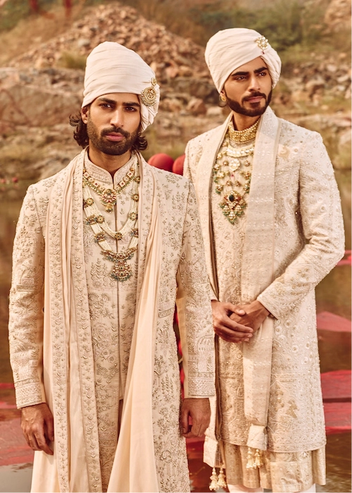 Two men in traditional embroidered sherwanis standing outdoors with a natural background.