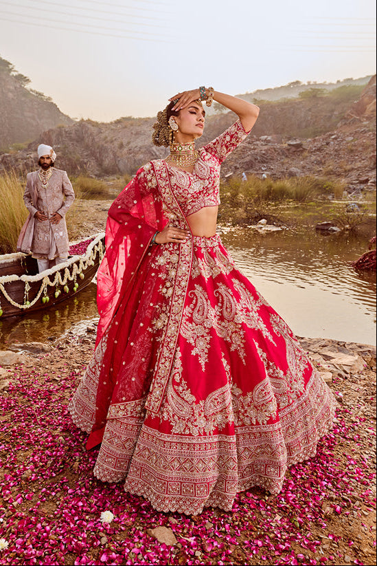 Woman in a red and gold traditional outfit standing by a lake with mountains in the background