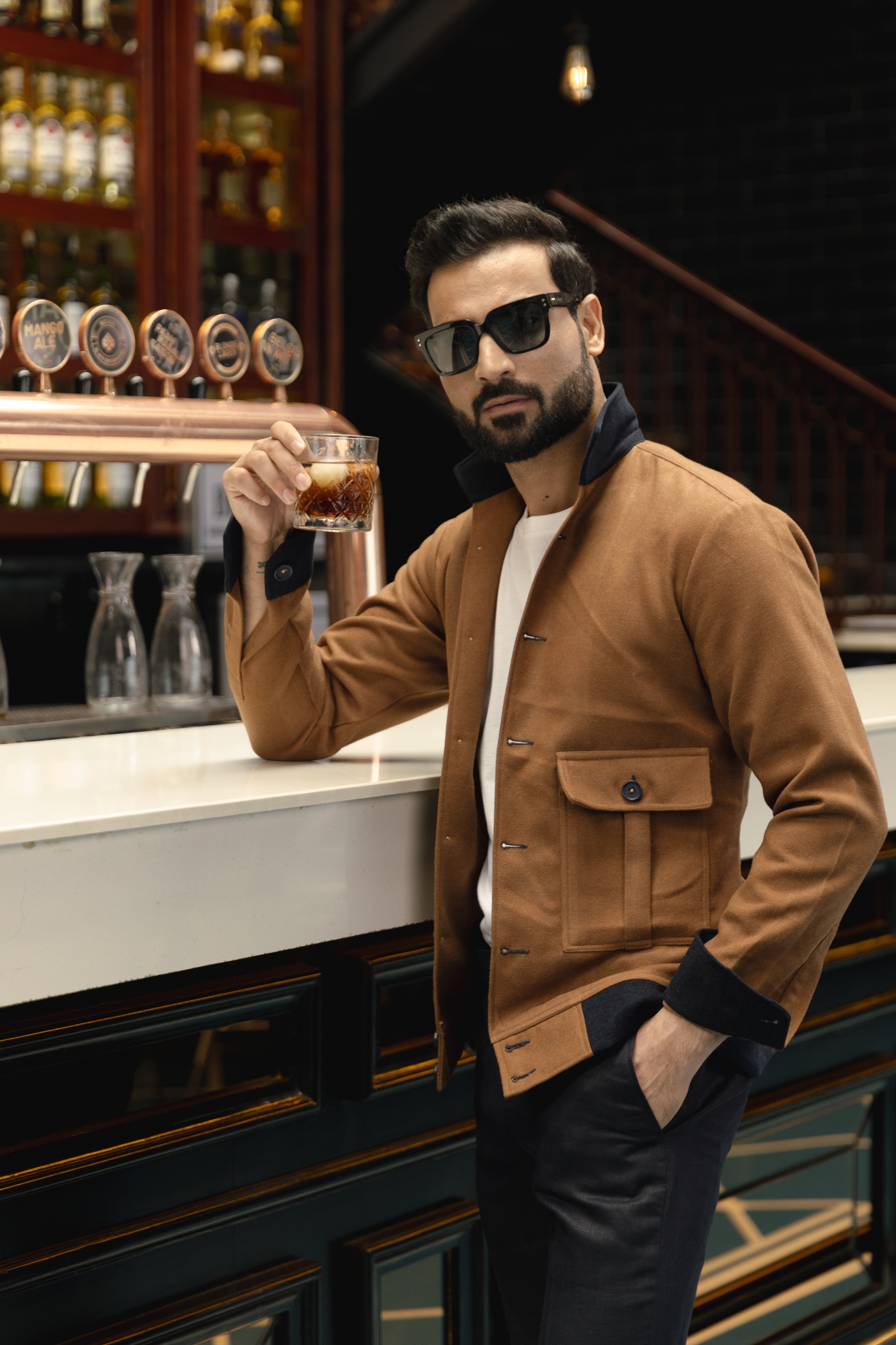 A man wearing a mustard yellow bomber jacket with patch pockets, sunglasses, and a white shirt, standing at a bar.