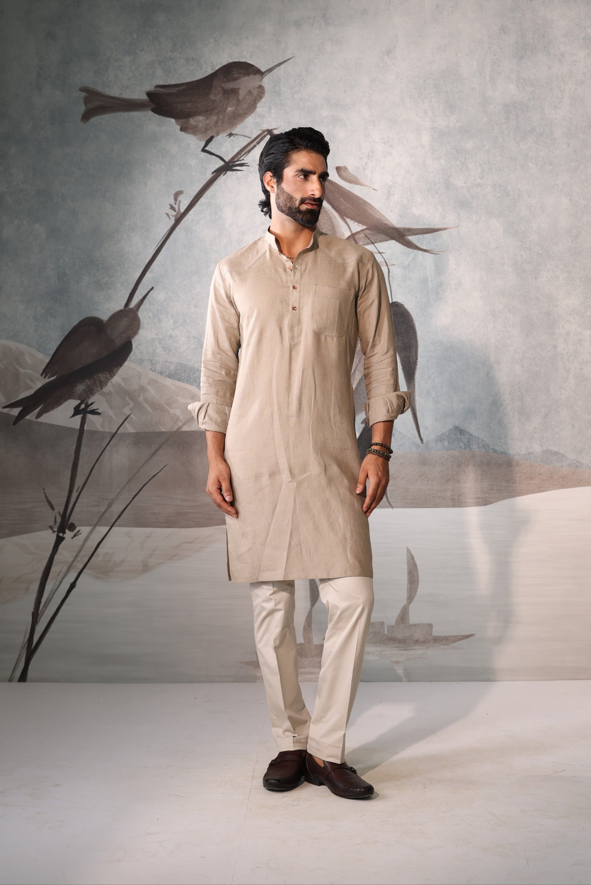 Man wearing a beige kurta with white pants standing in front of a wall with bird artwork.