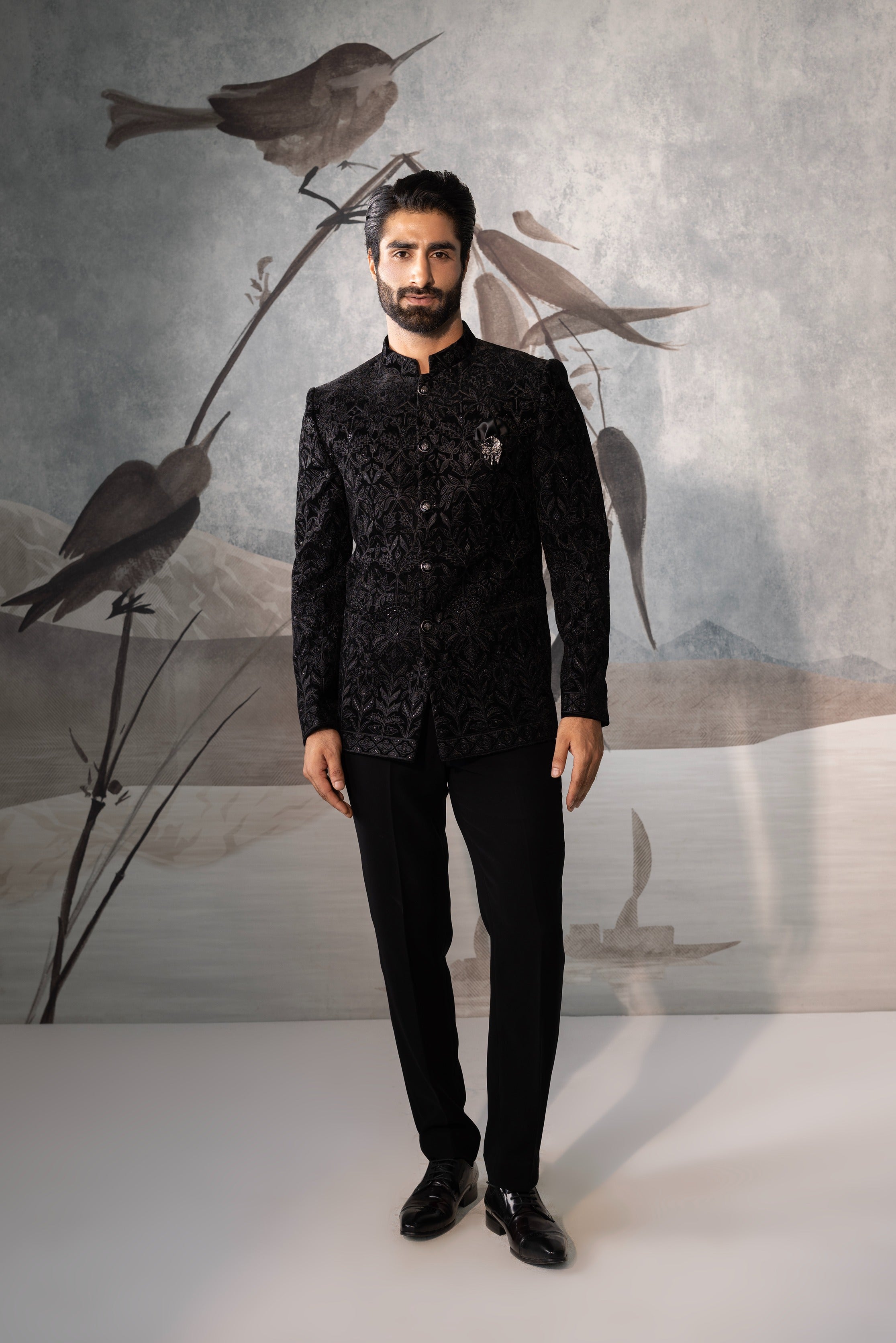 Man wearing a black traditional outfit with a decorative pattern against a background of stylized birds.