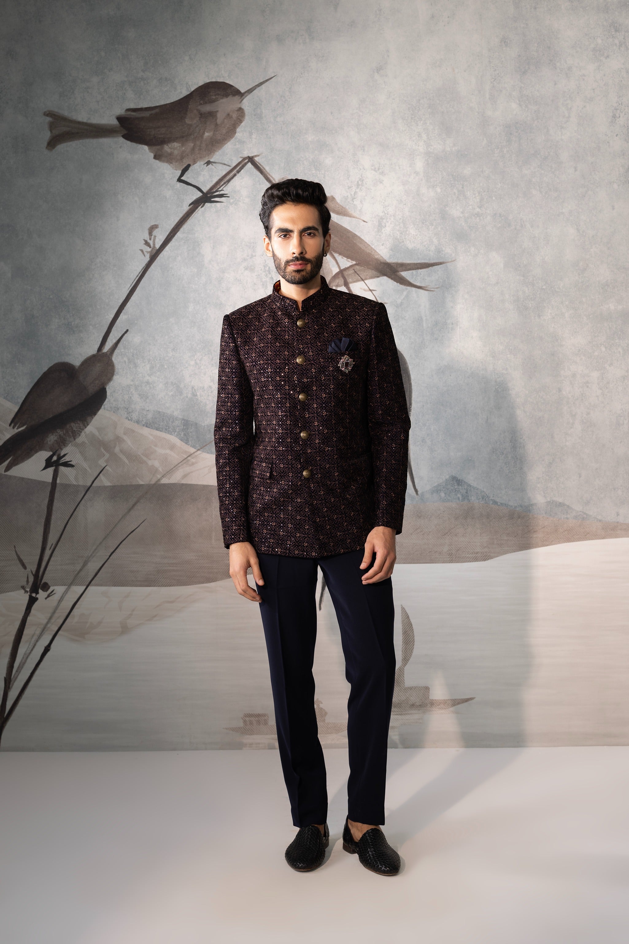 Man wearing a dark patterned jacket and pants standing in front of a wall with bird decorations.