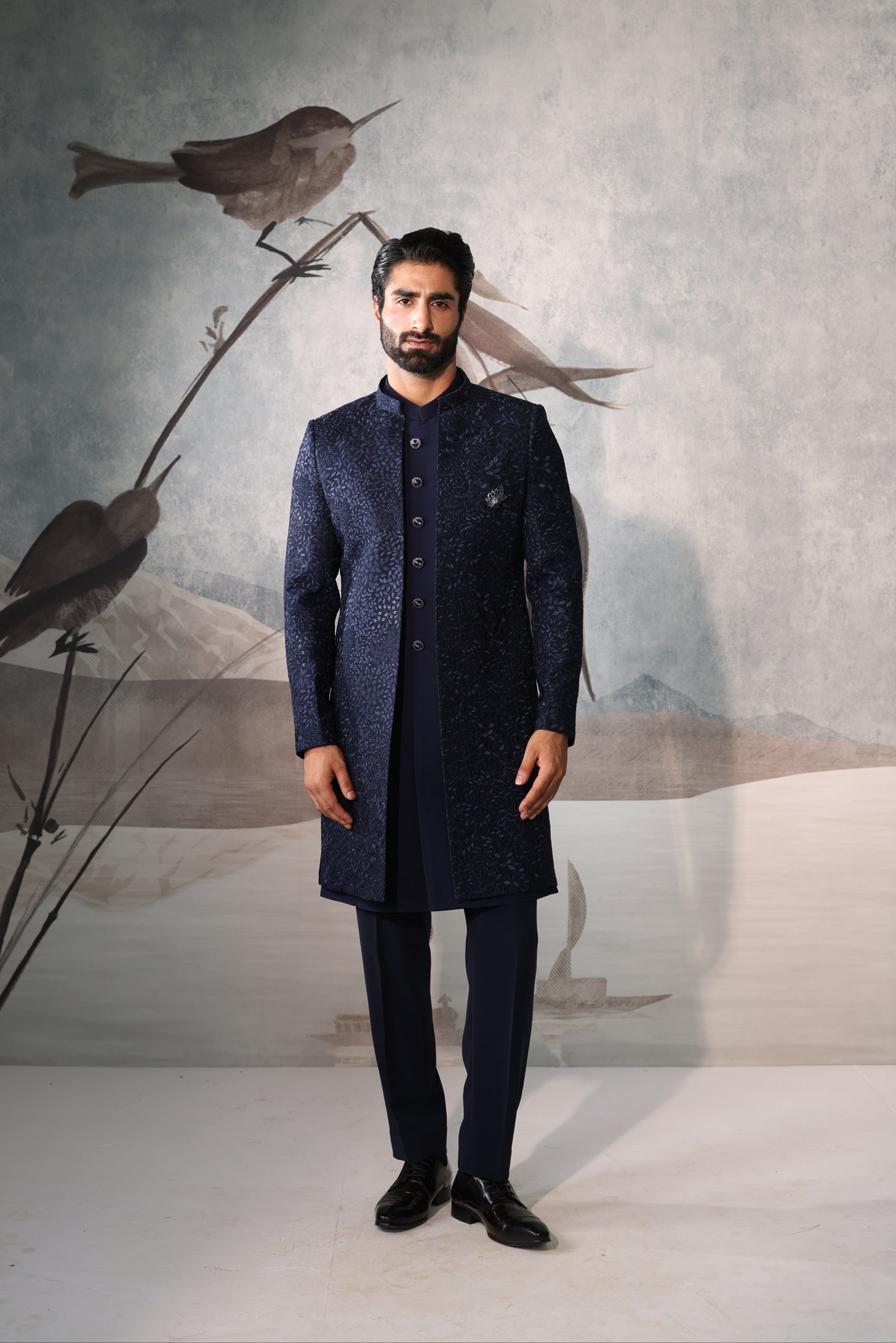 Man wearing a dark blue traditional outfit standing in front of a wall with bird illustrations.