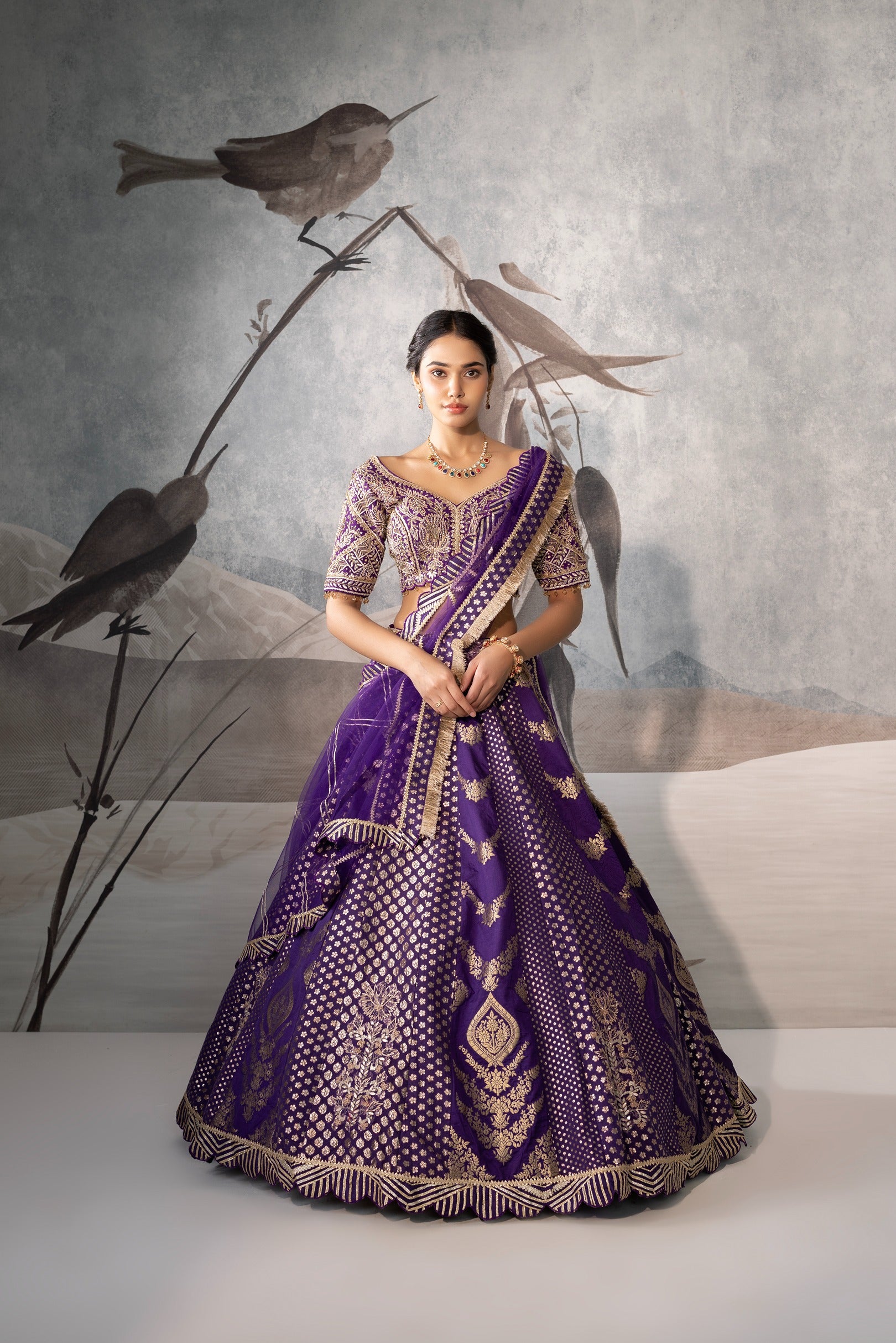 Woman in a purple traditional outfit standing against a neutral background with decorative birds.