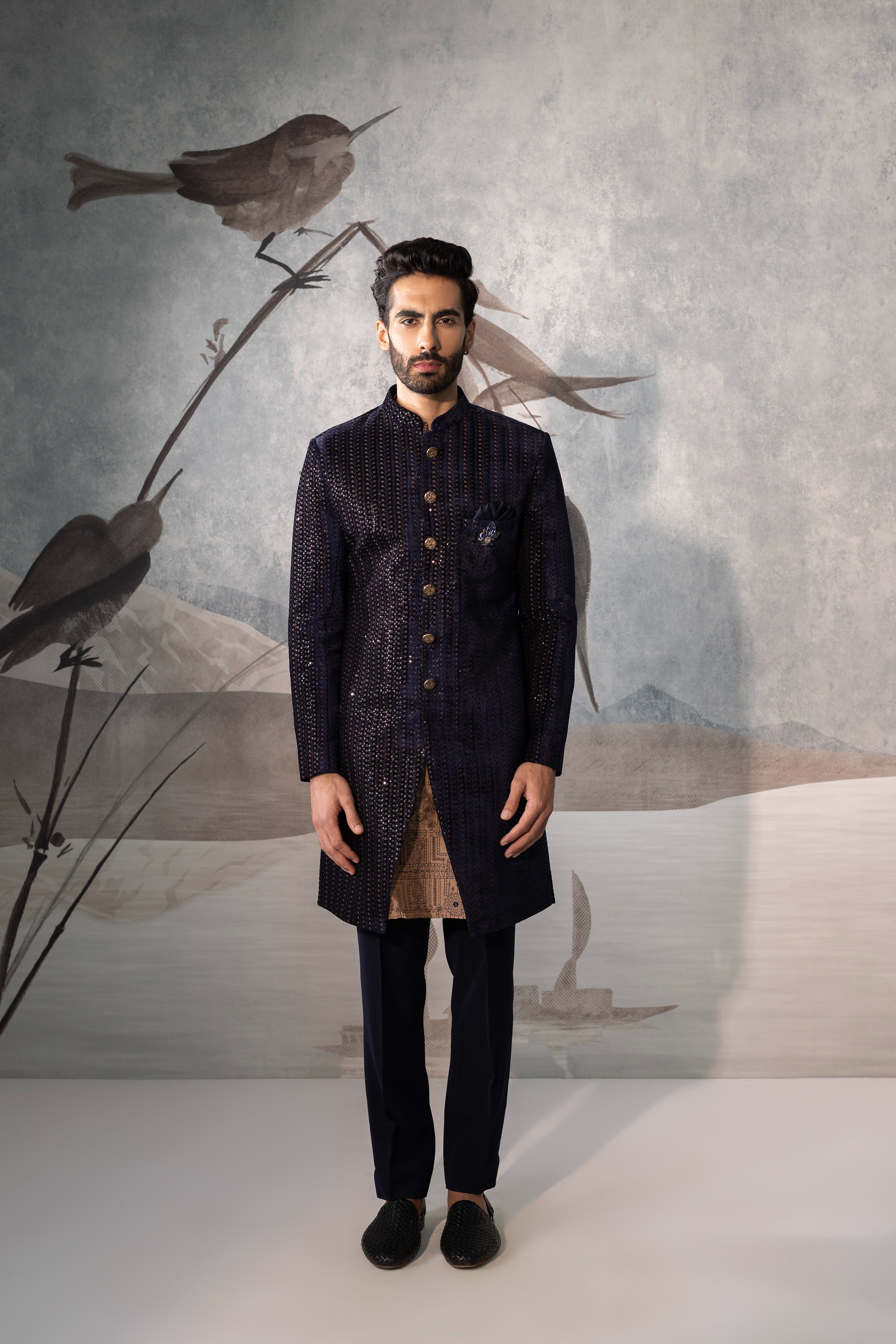 Man wearing a dark traditional outfit with a stylized bird illustration on the wall behind him.