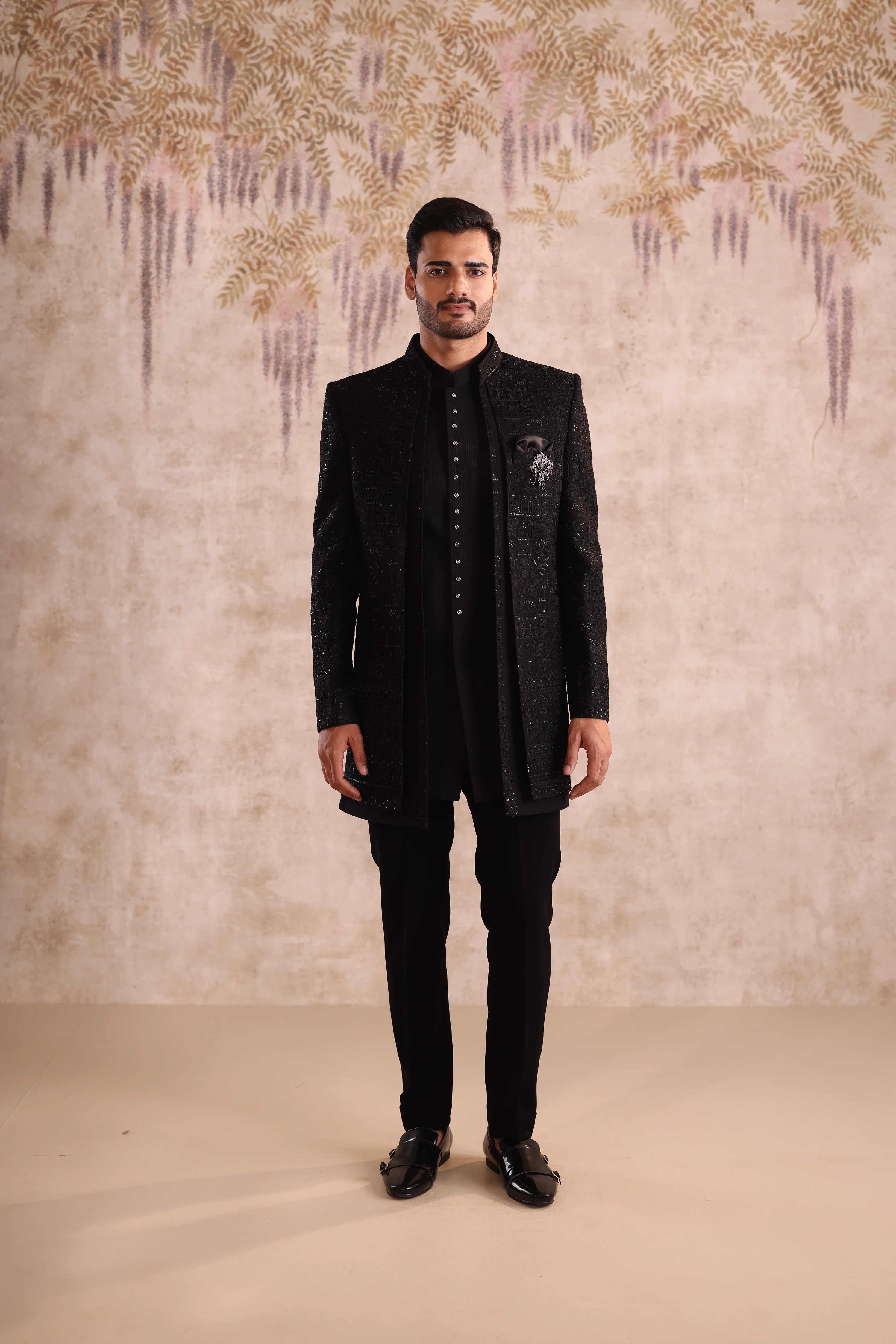 Man wearing a black traditional outfit against a decorative wall.