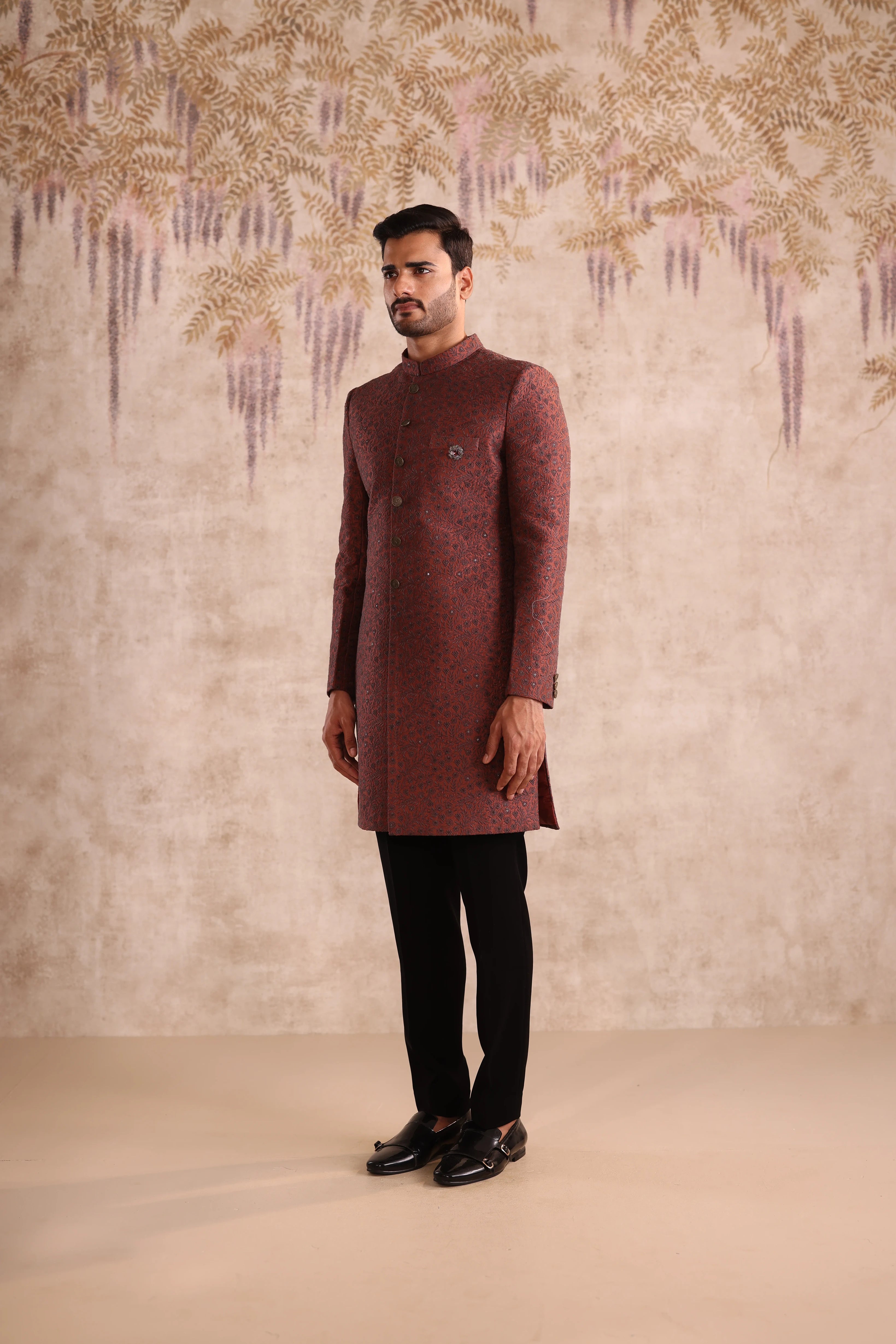 Man wearing a maroon kurta with black pants against a decorative wall.