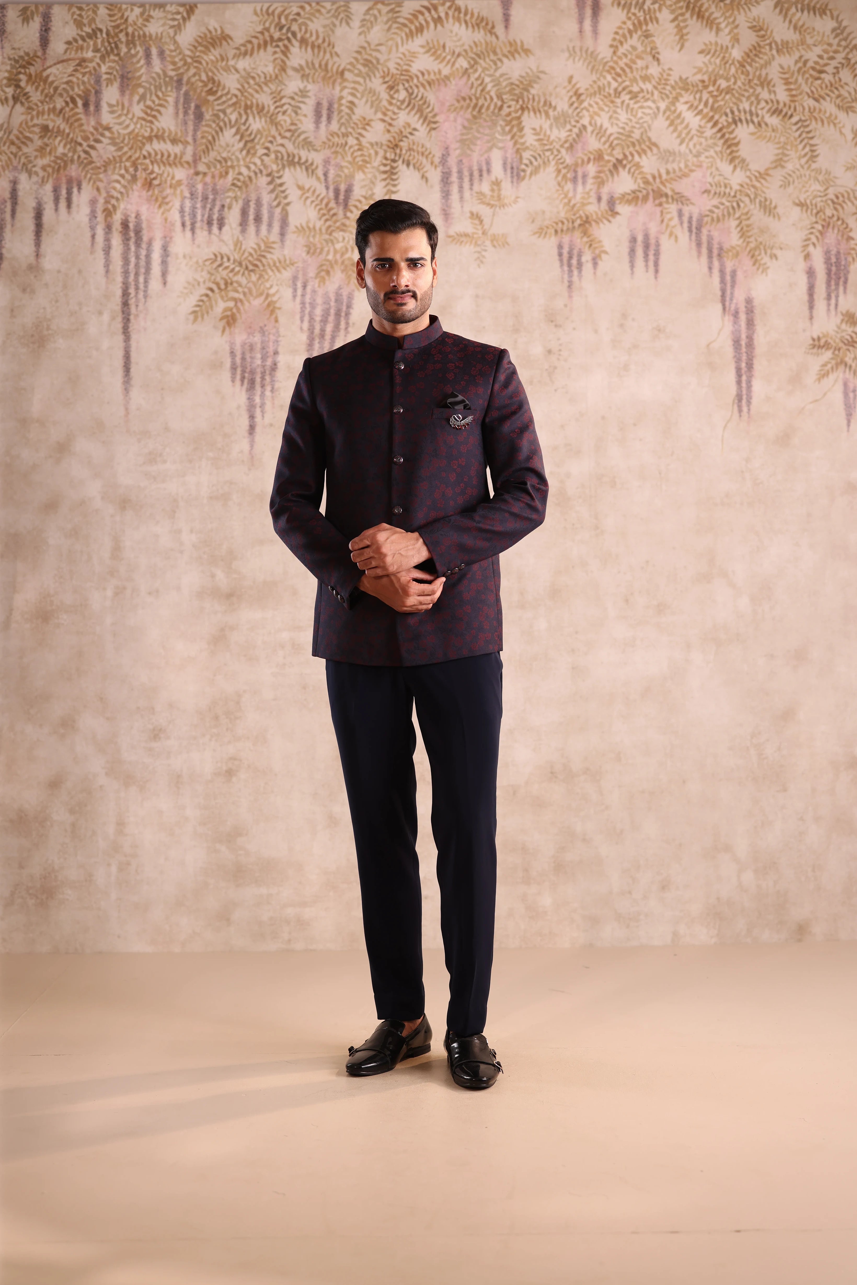 Man wearing a dark purple traditional outfit against a decorative wall.