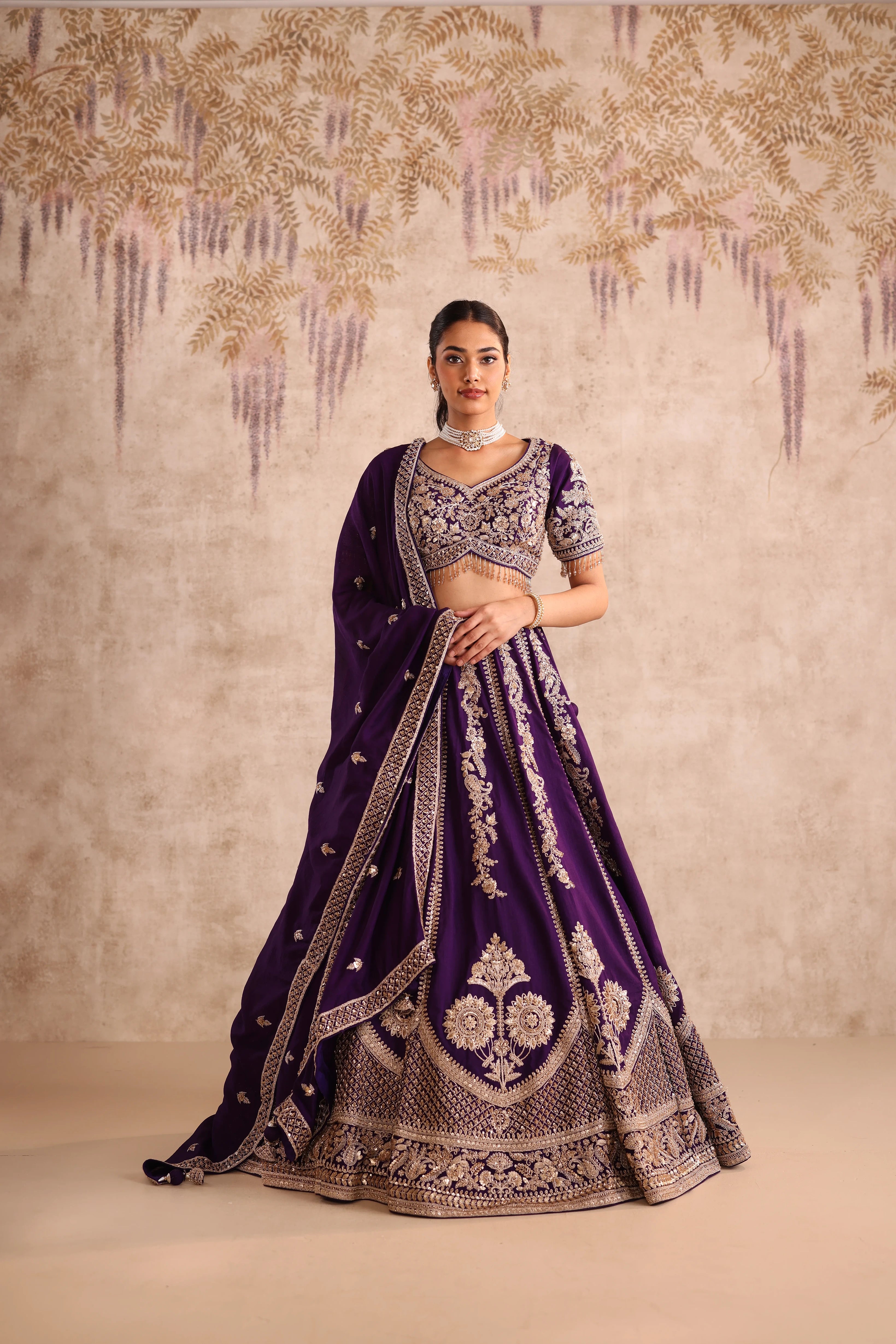 Woman wearing a purple embroidered traditional outfit against a decorative wall.
