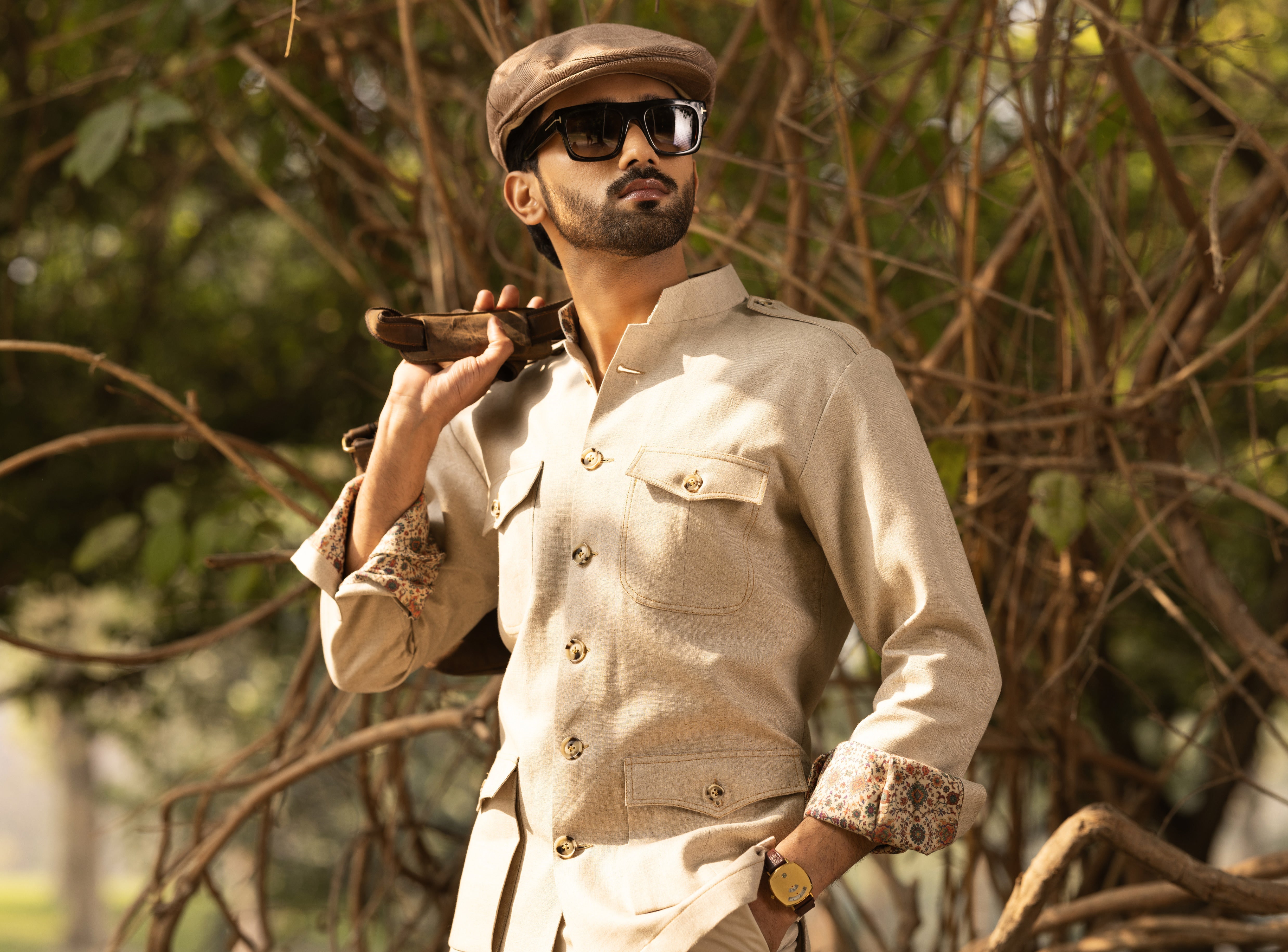 Man in beige safari outfit with hat and sunglasses, holding a rifle, against a natural background.