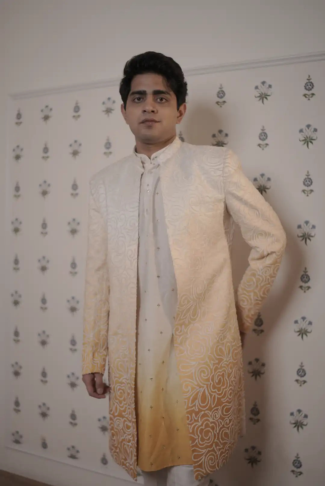 Man wearing a traditional cream sherwani with gold patterns against a decorative wall.