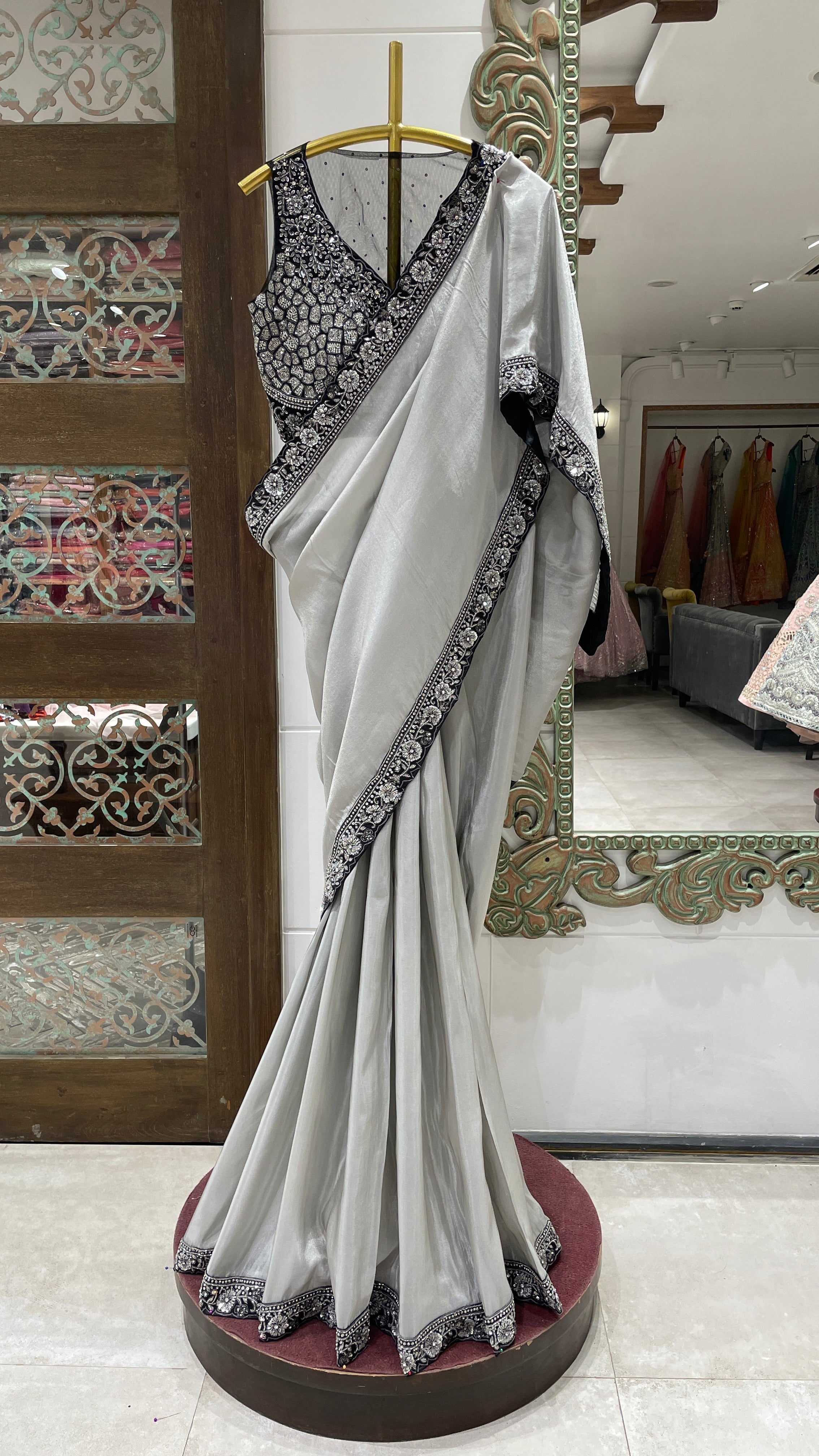 A silver tissue saree with black border displayed on a mannequin in a store with traditional attire and decorative patterns in the background.