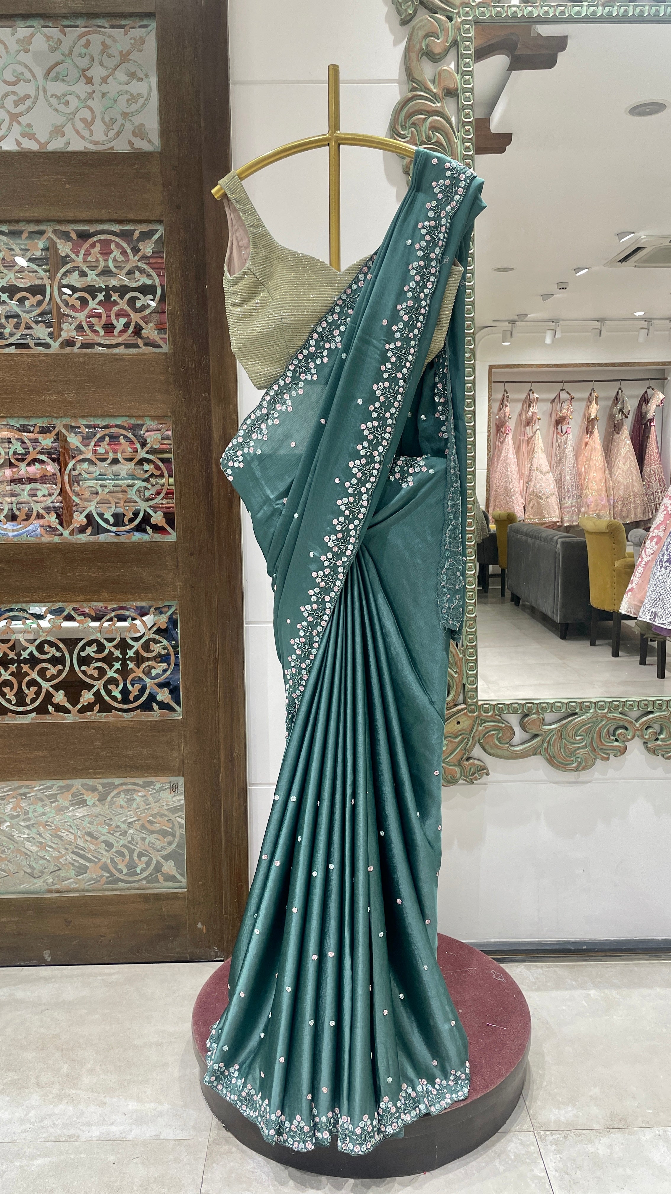 A green crepe saree with floral motifs displayed on a mannequin in a store with intricate floral patterns and a red base.