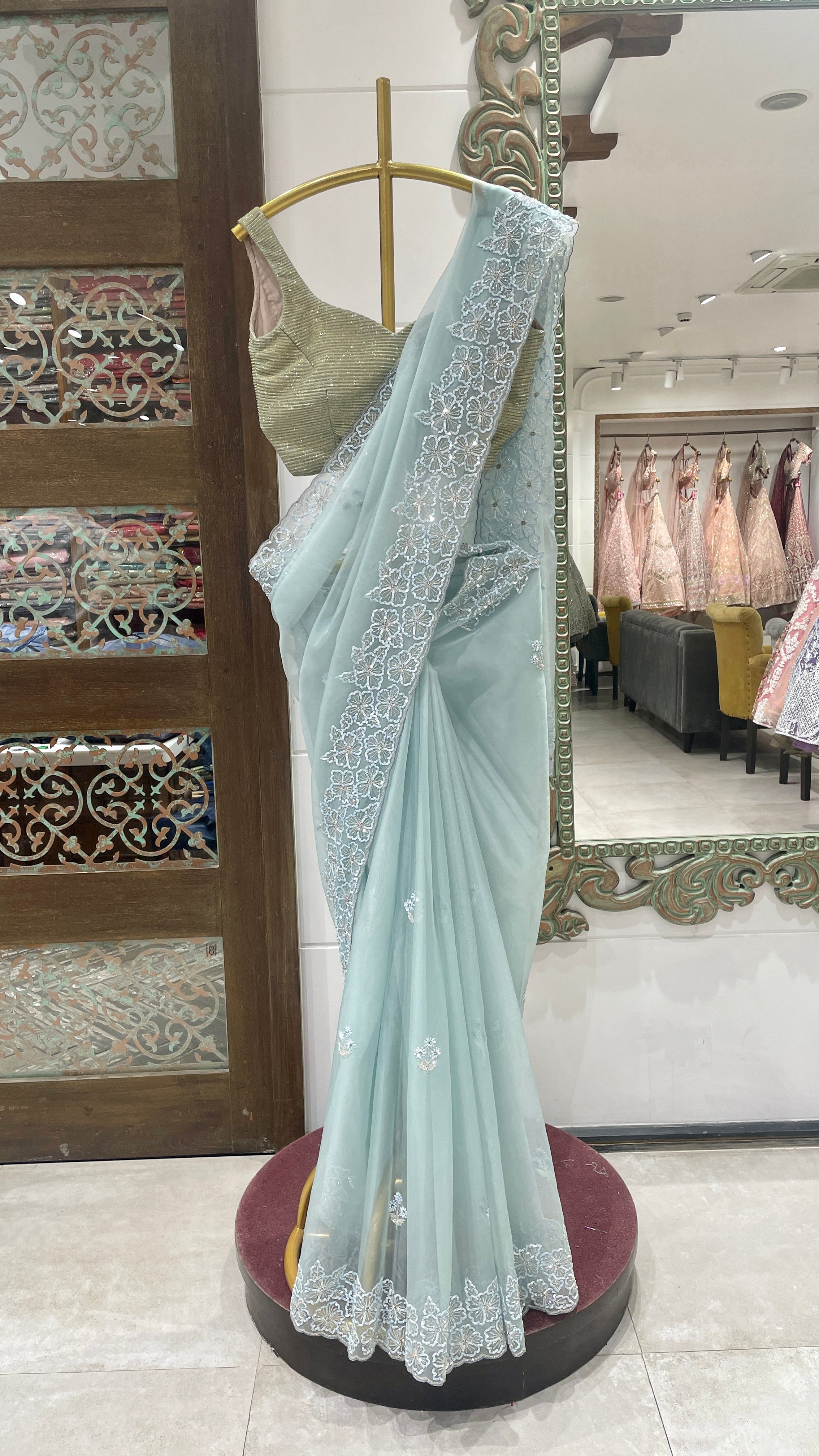 A pastel blue organza saree with embellishments displayed on a mannequin, with the saree's end showing detailed floral pattern and embellishments.