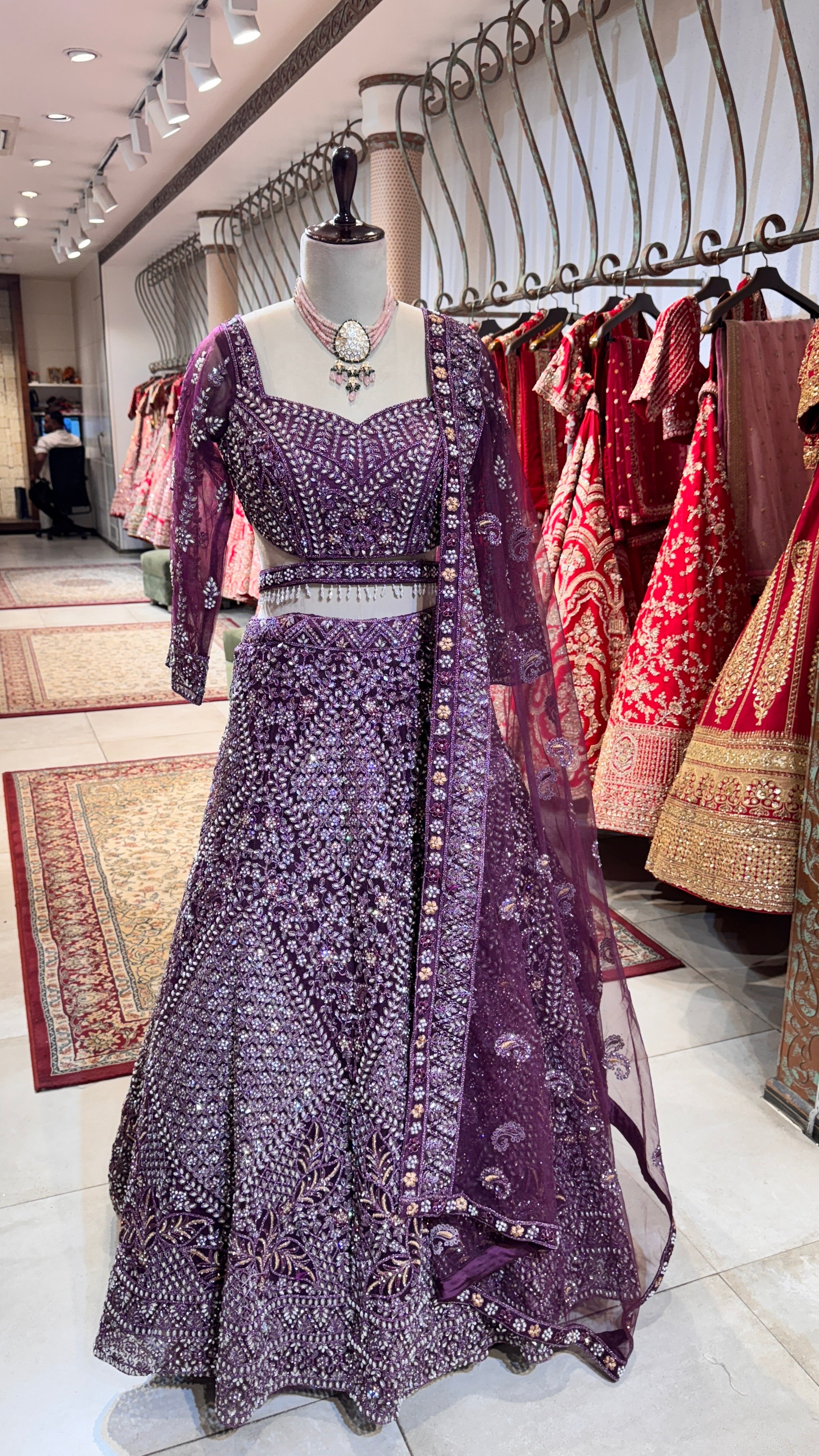 A mannequin displaying a rich purple lehenga adorned with intricate embroidery and a matching blouse, paired with a sheer dupatta with embroidered borders.