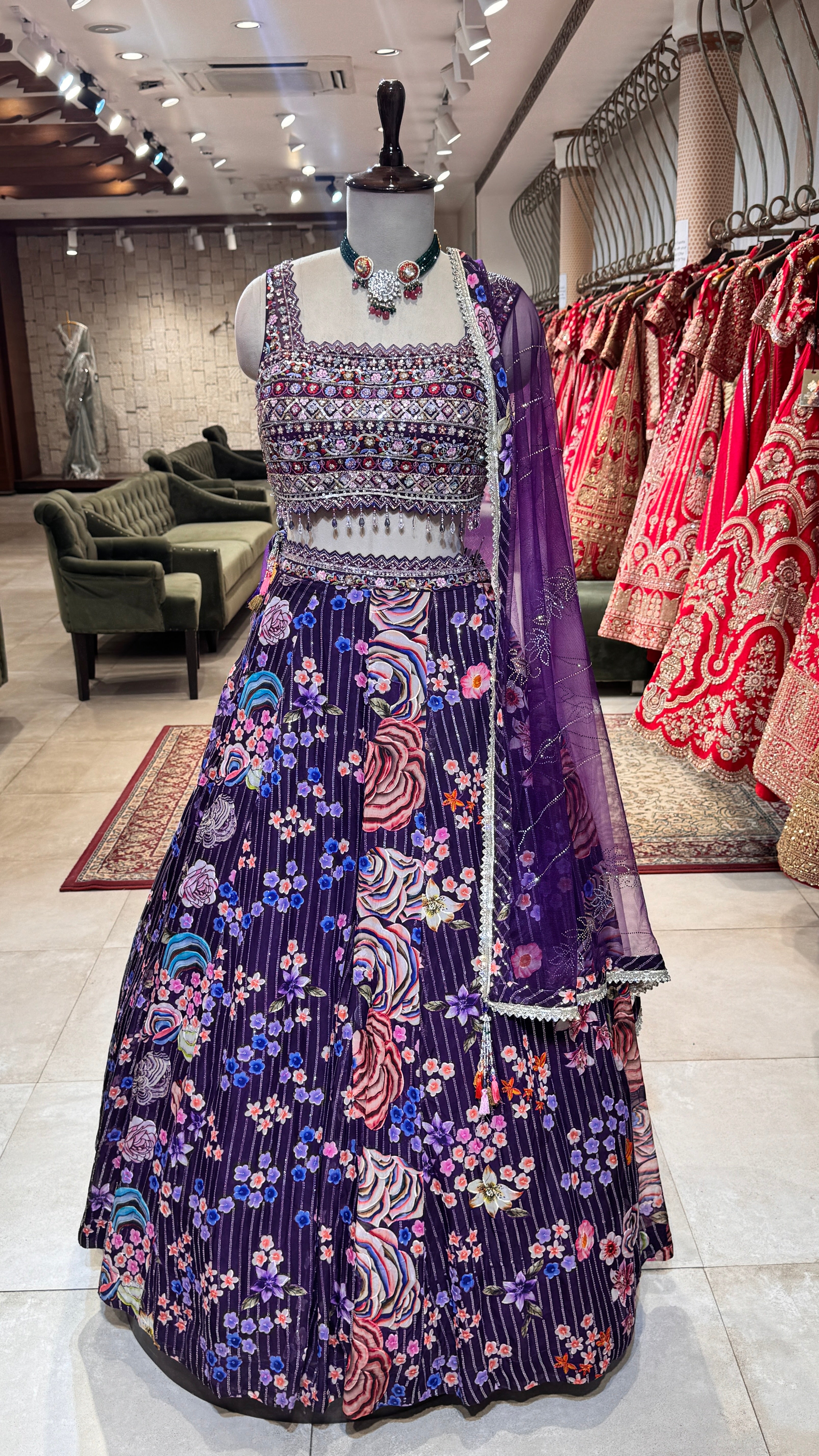 A mannequin displaying a purple floral lehenga with a blouse and dupatta, the ensemble is crafted with floral embroidery and border accents.