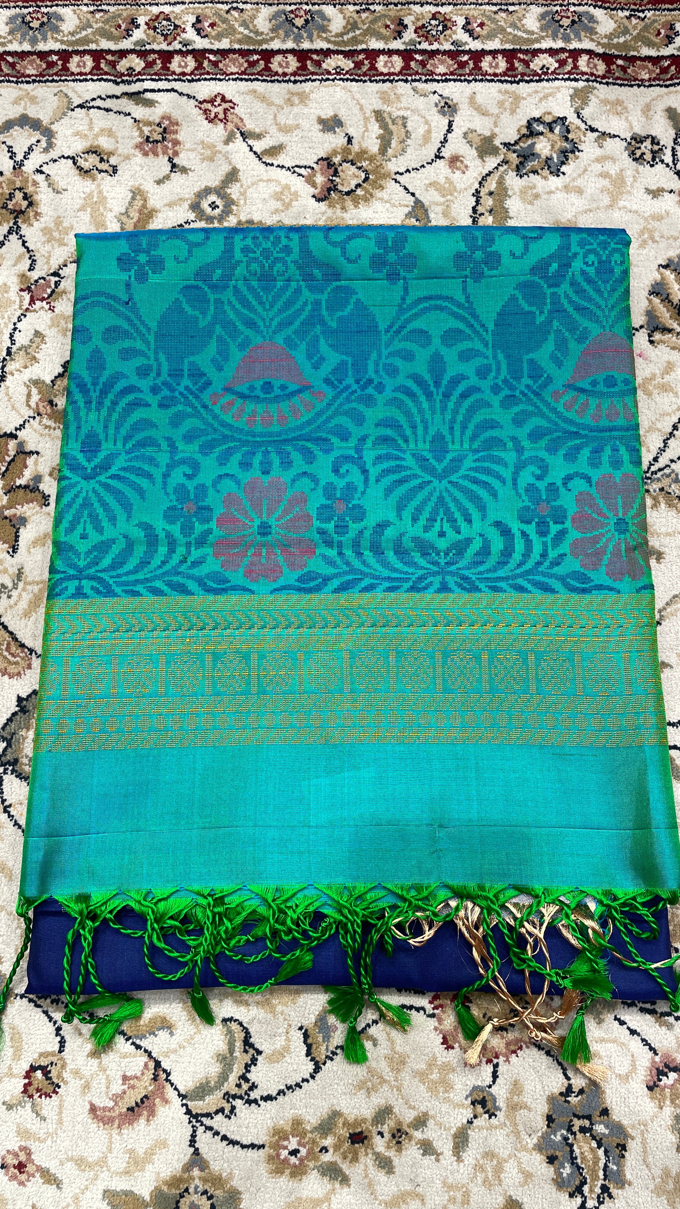 A folded turquoise blue handloom silk saree with intricate floral designs and a green border, displayed on a floral patterned fabric.