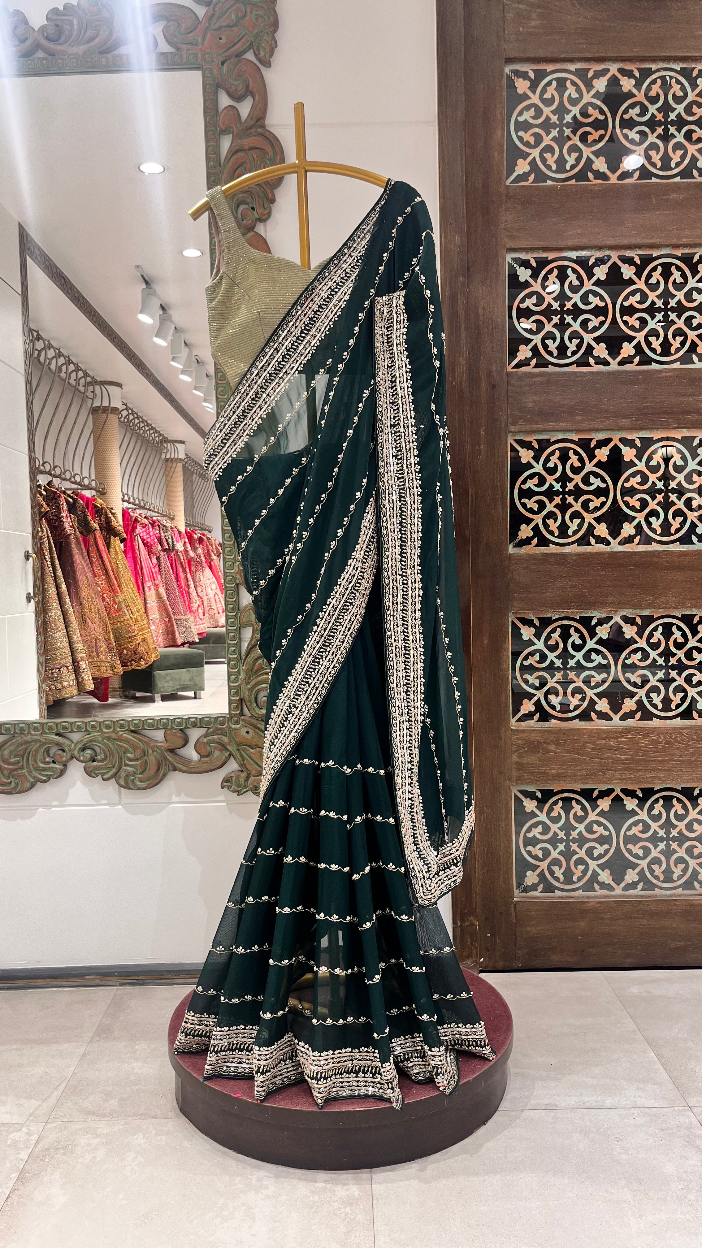 A green saree with gold embellishments displayed on a mannequin in a store with traditional Indian attire and decorative patterns in the background.
