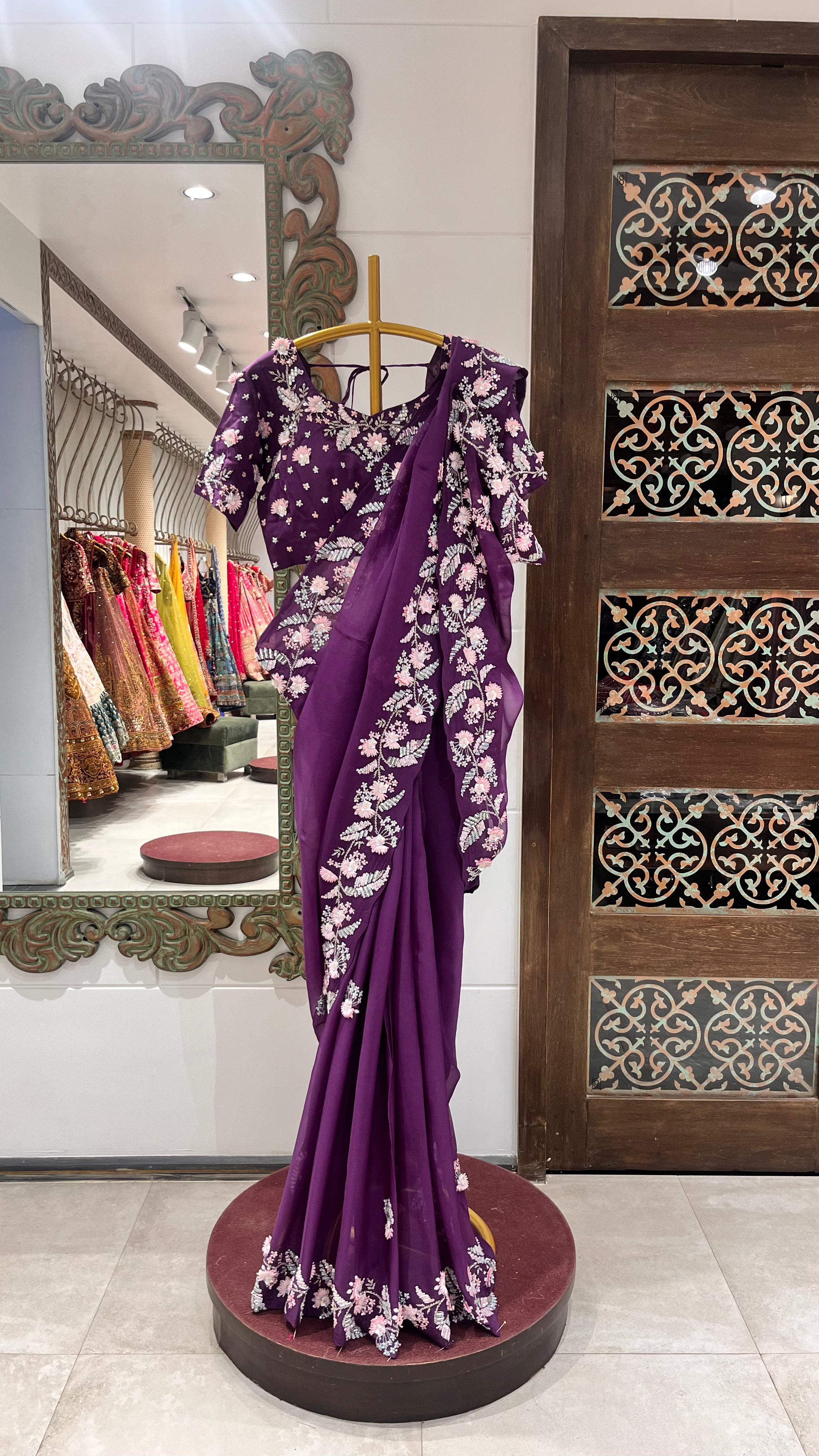 A purple organza saree with floral motifs displayed on a mannequin in a store with various other sarees in the background.