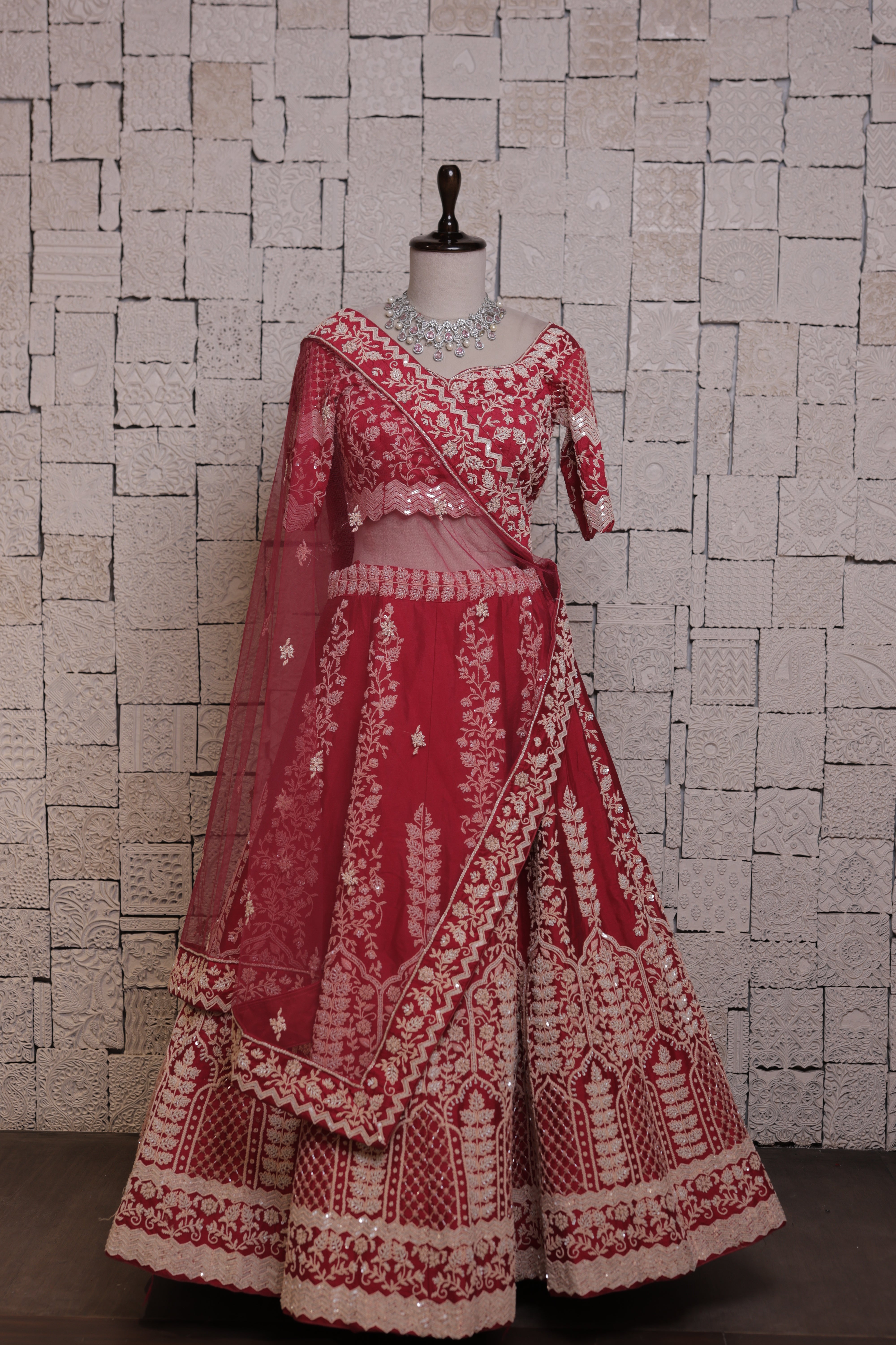 A red bridal lehenga with intricate flower pot embroidery and zari & sequin work, displayed on a mannequin against a brick wall backdrop.