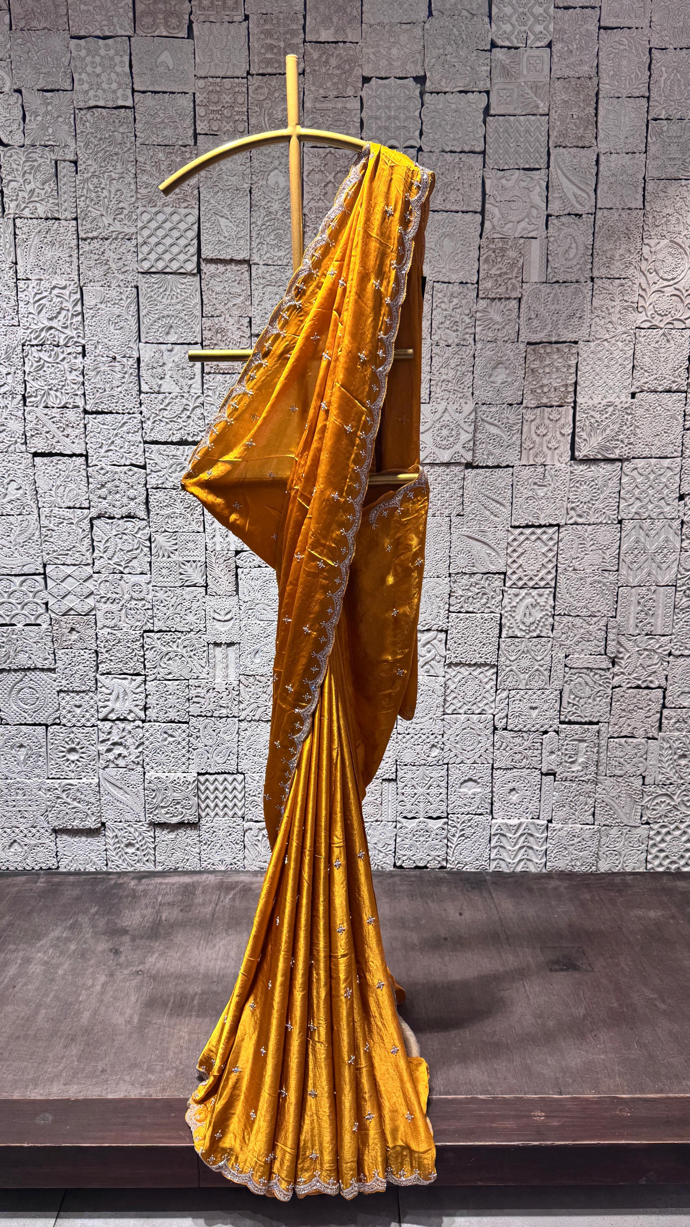 A mustard yellow saree with a zig-zag border displayed on a hanger against a brick wall background.