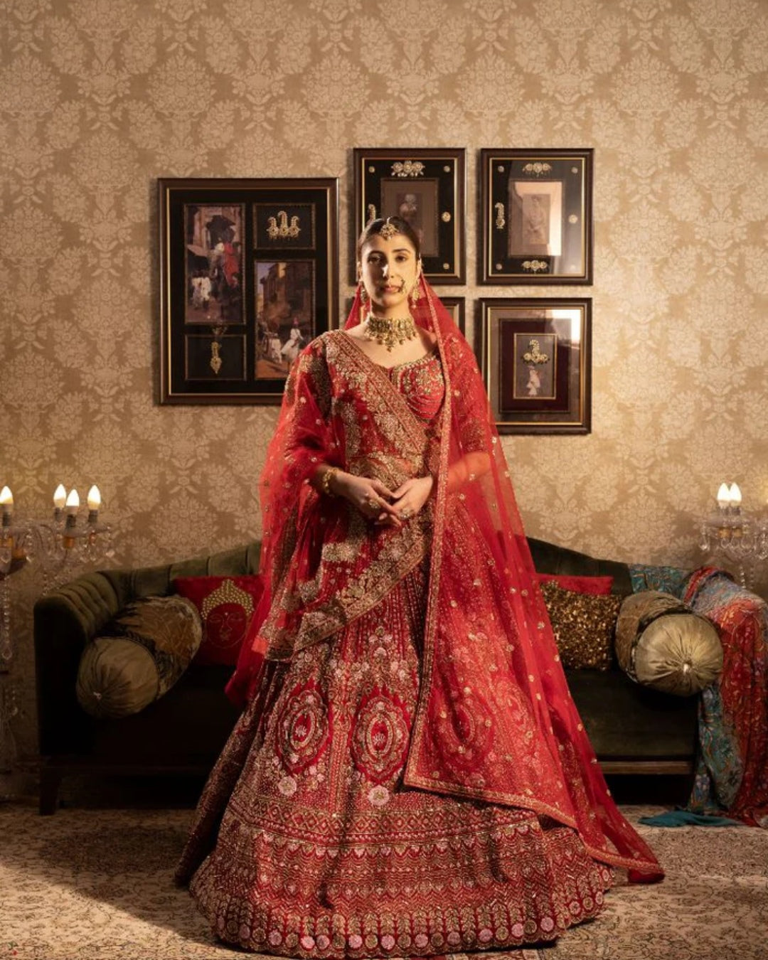 Woman in a red and gold traditional outfit standing in a decorated room with framed pictures on the wall.