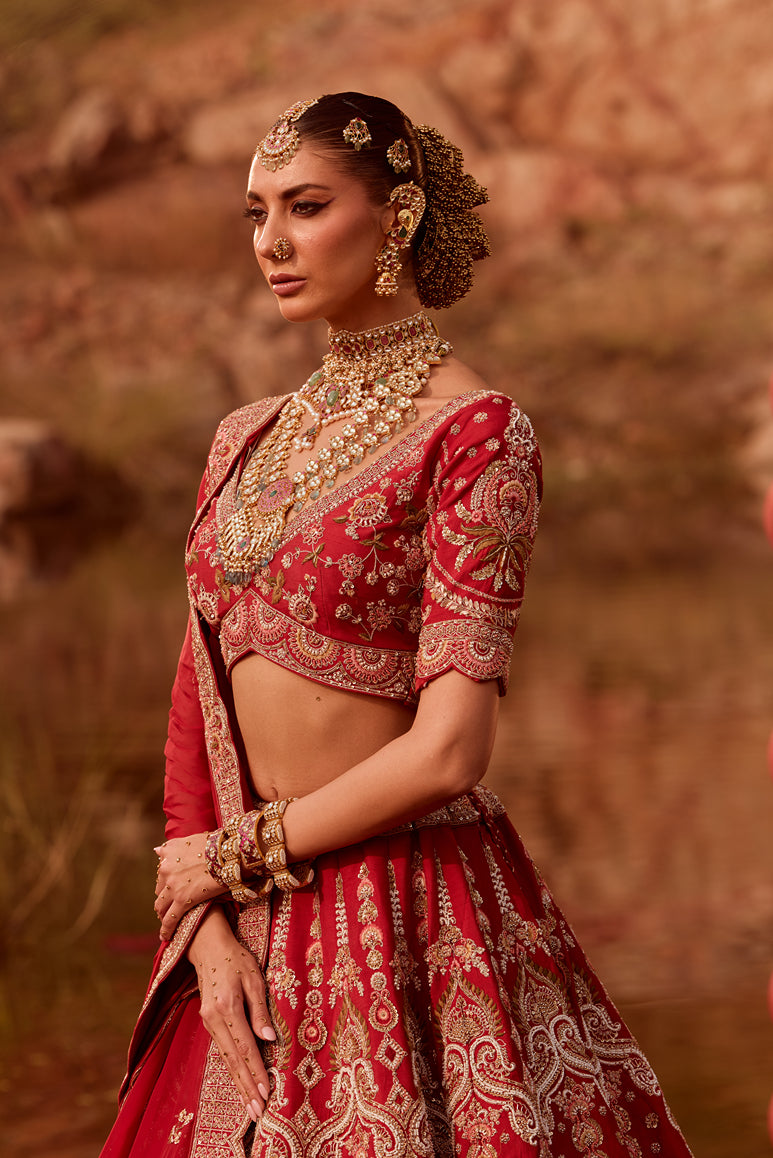 Woman in traditional red and gold embroidered outfit with jewelry against a blurred natural background