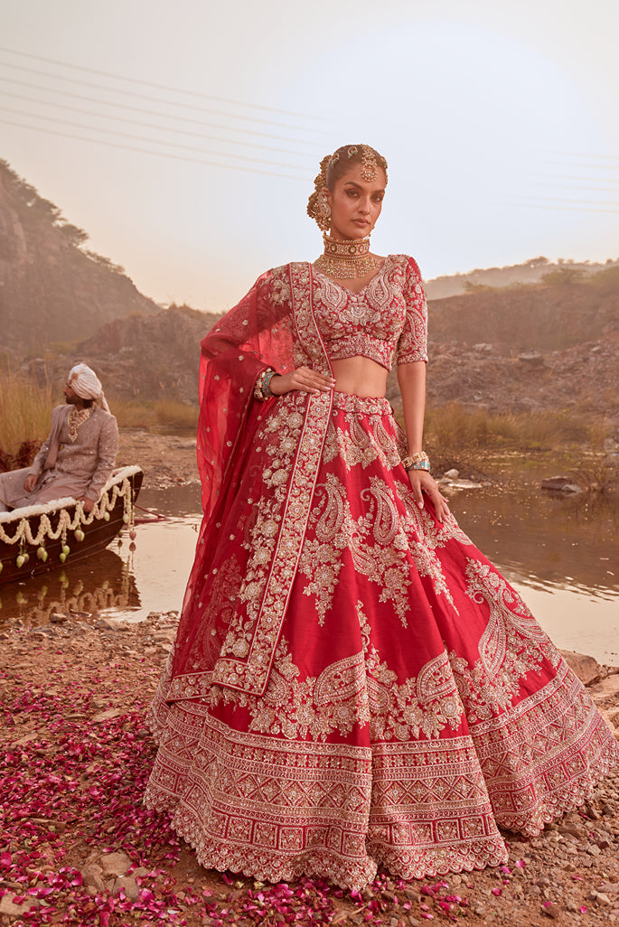 Woman in a red and gold traditional outfit standing by a lake with another person in the background.