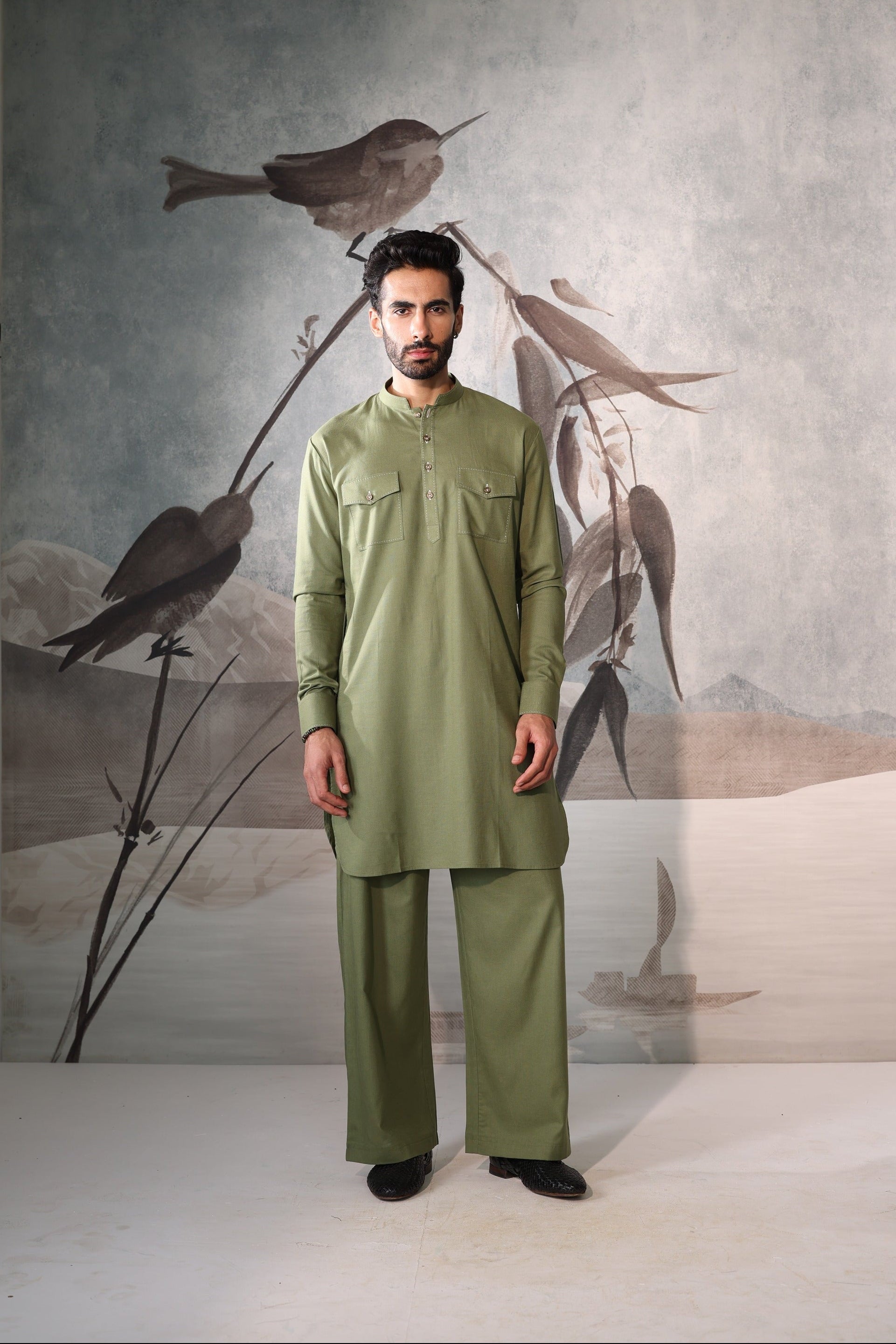 Man wearing a green traditional outfit standing in front of a wall with bird artwork.