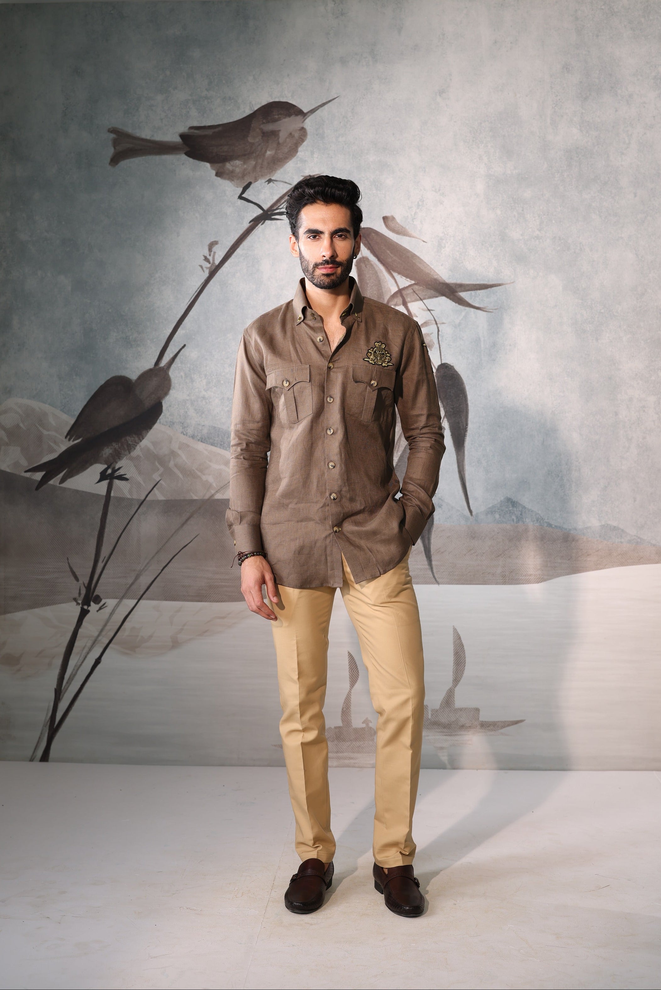 Man wearing a brown shirt and beige pants standing in front of a wall with bird artwork.