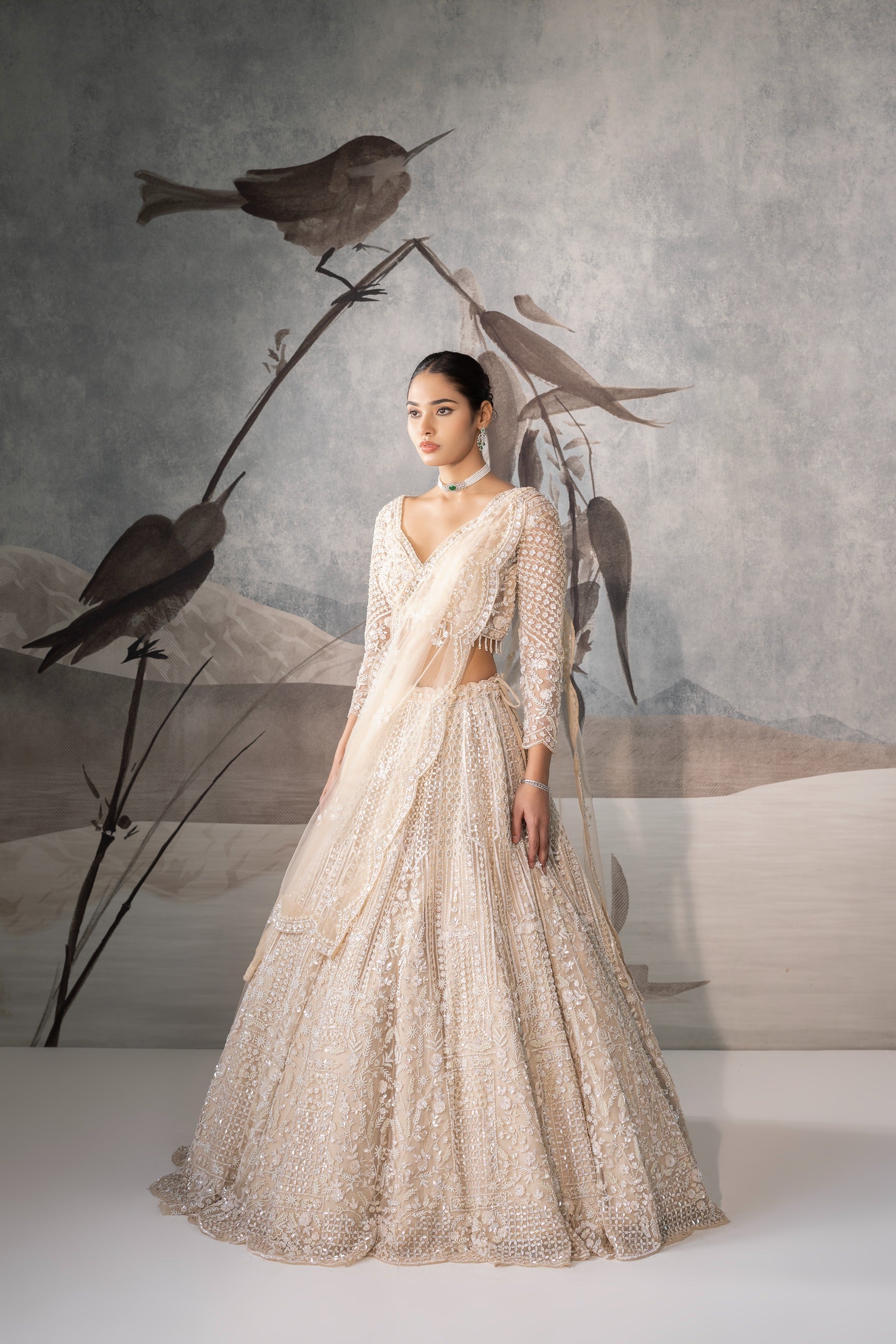 Woman in a beige embroidered dress standing in front of a decorative background with birds.