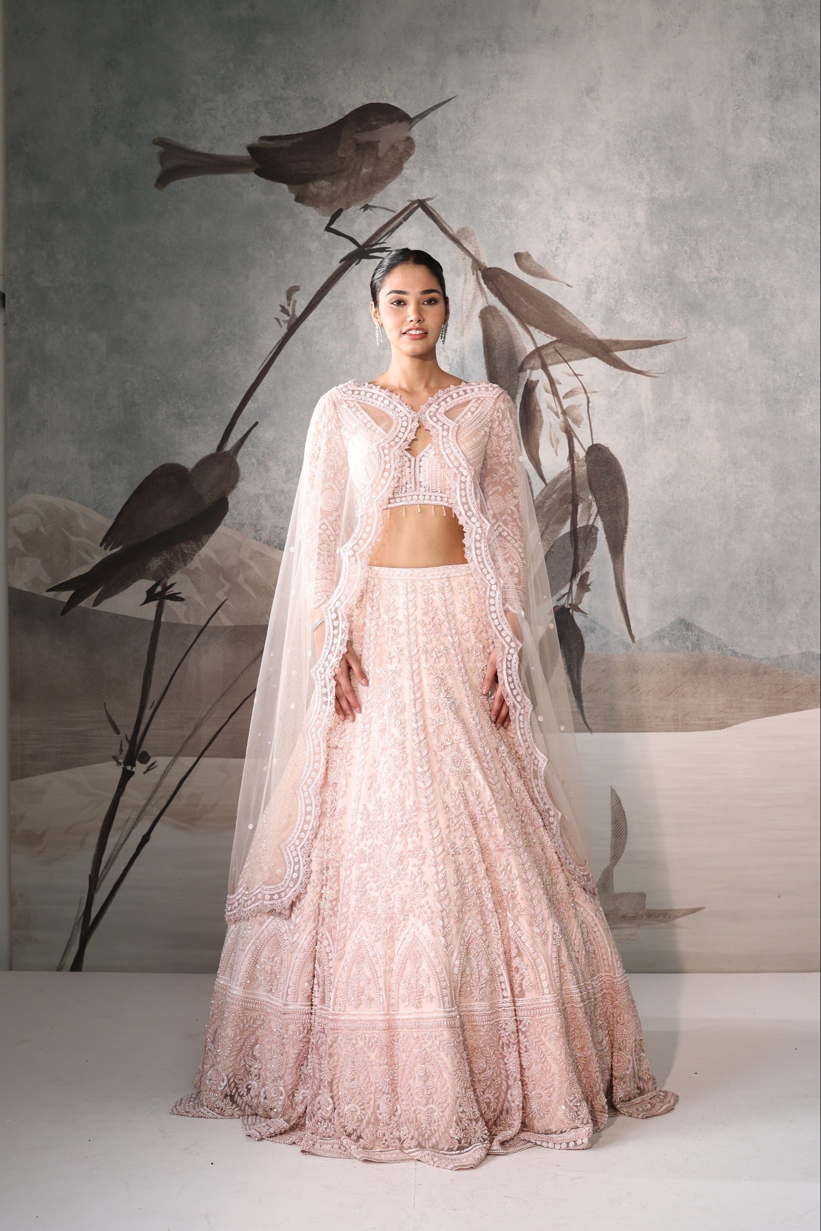 Woman in a pink traditional outfit standing in front of a decorative wall with birds.