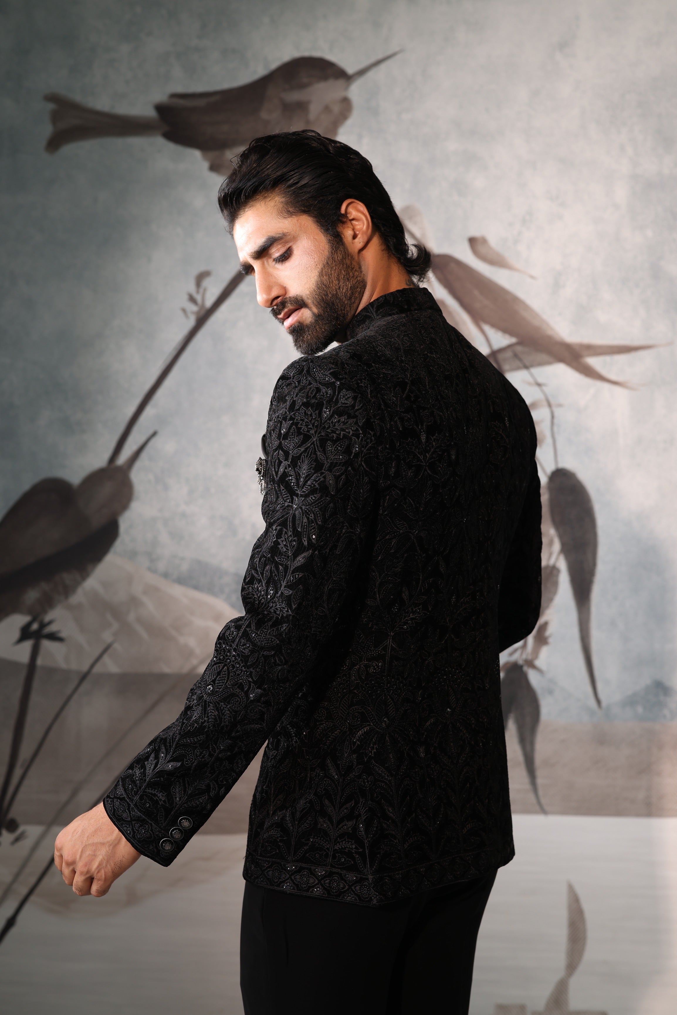 Man wearing a black textured jacket in front of a decorative wall with bird illustrations.