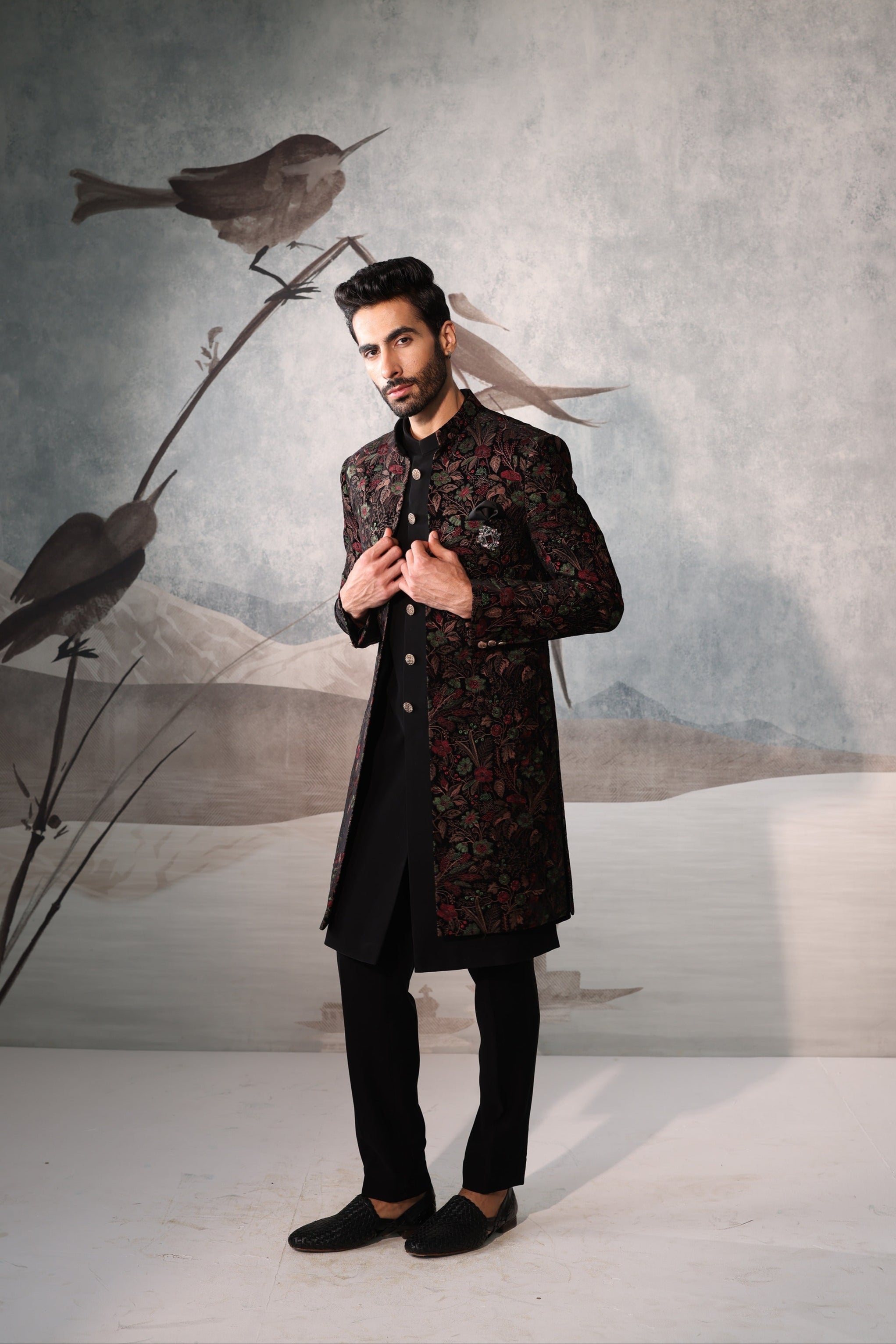 Man in a dark traditional outfit standing in front of a wall with bird illustrations.