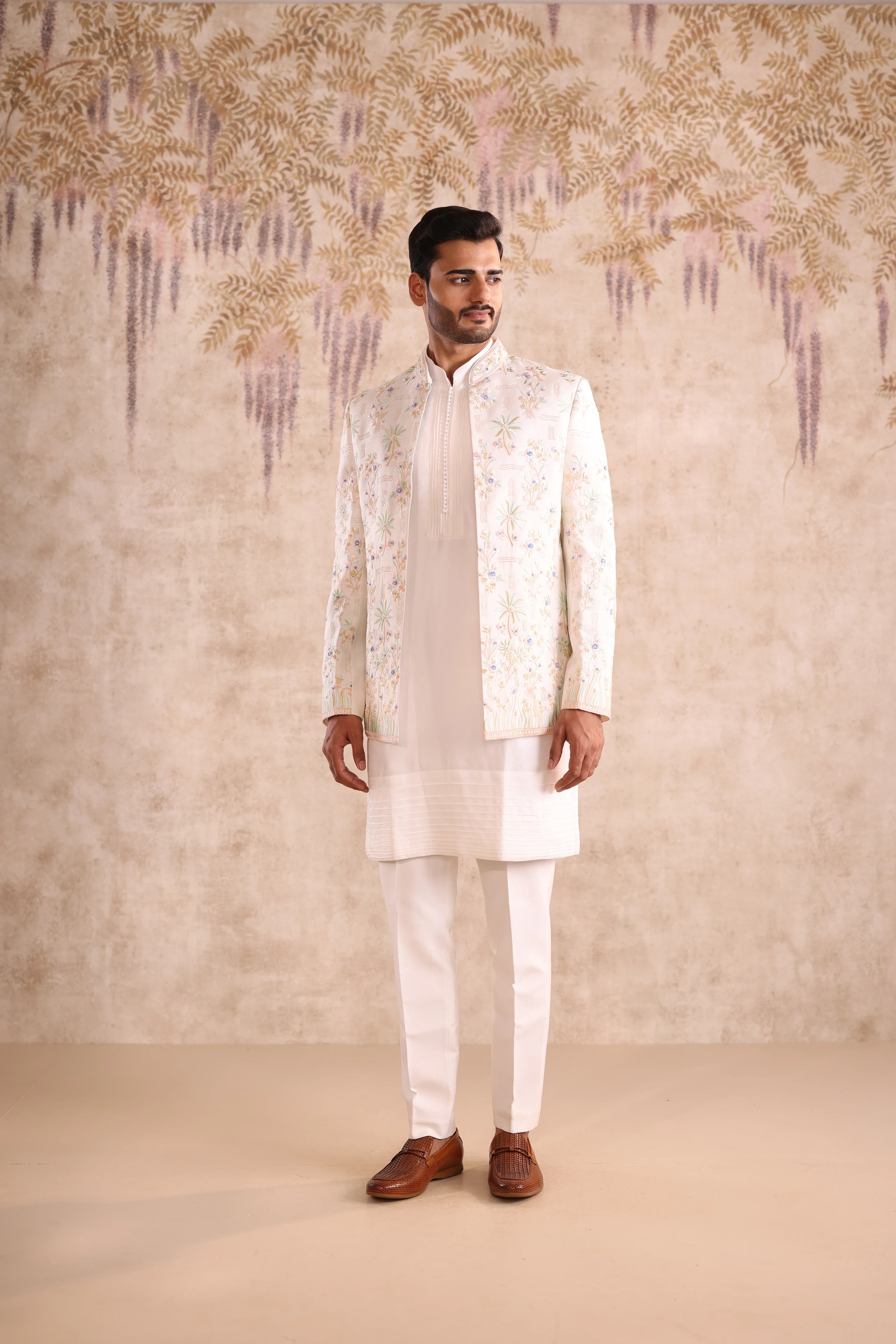 Man wearing a white embroidered sherwani against a decorative wall.