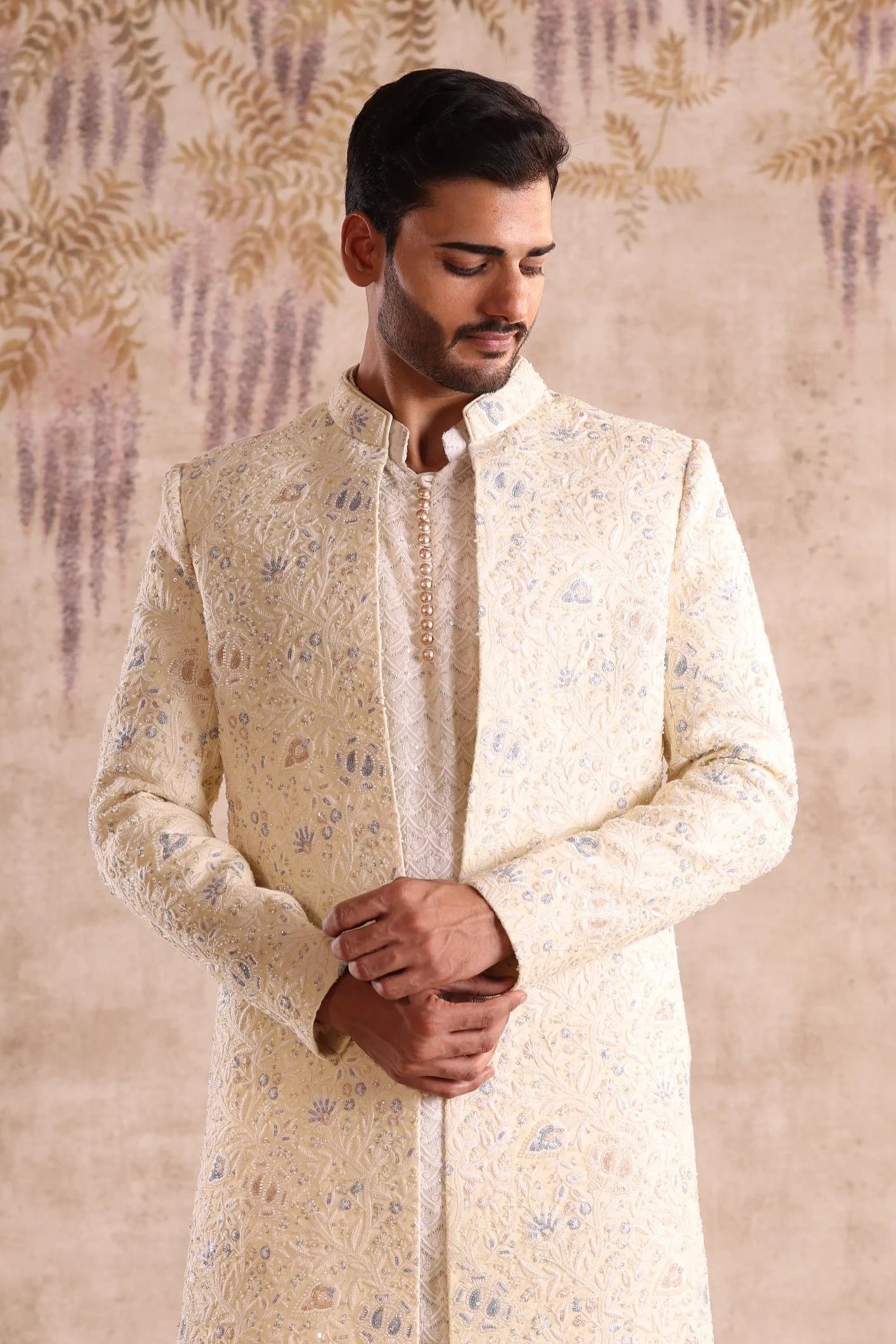 Man wearing a traditional cream sherwani with white pants against a decorative wall.