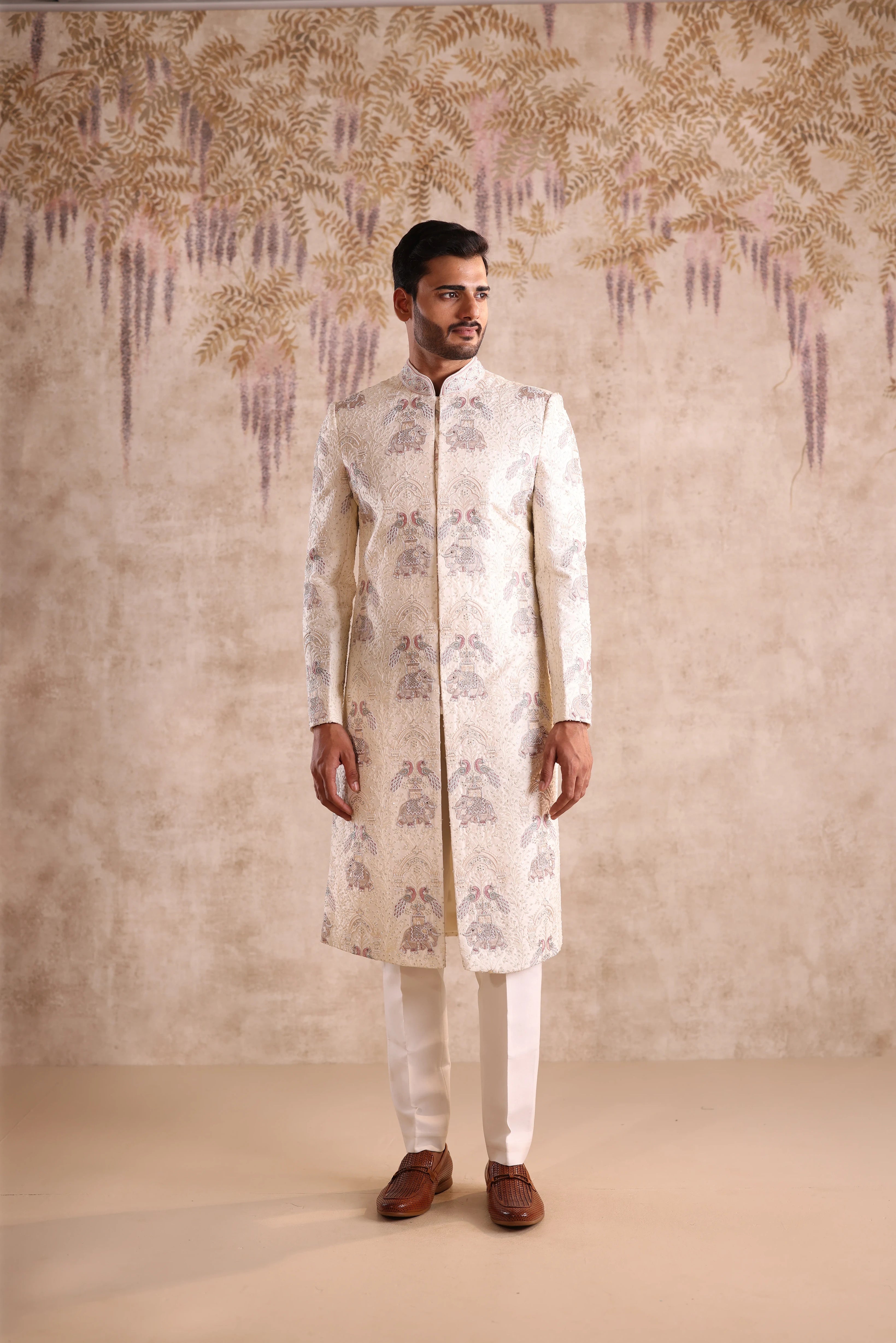 Man wearing a white sherwani with intricate patterns against a decorative wall.