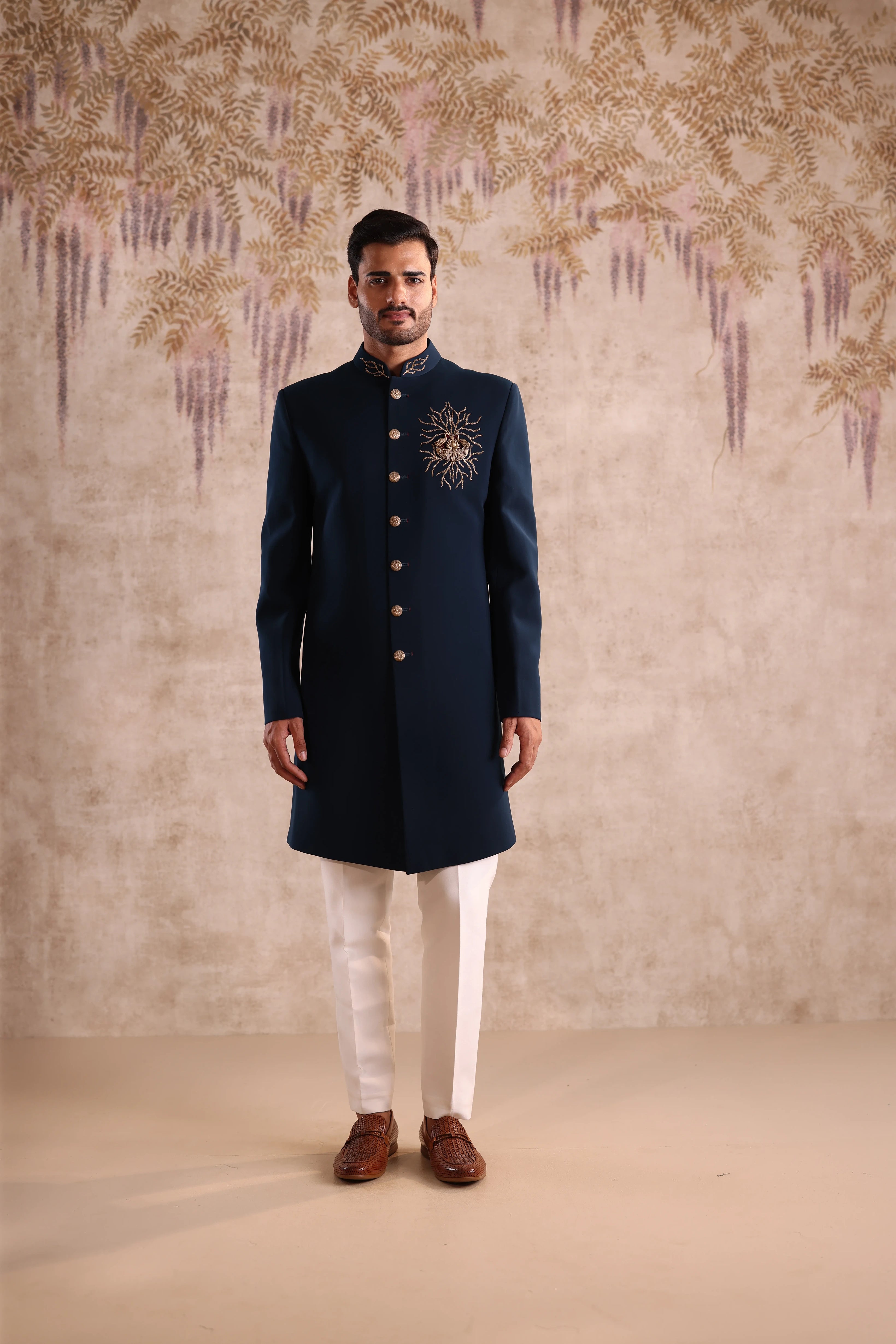 Man wearing a navy blue sherwani with white pants against a decorative wall.