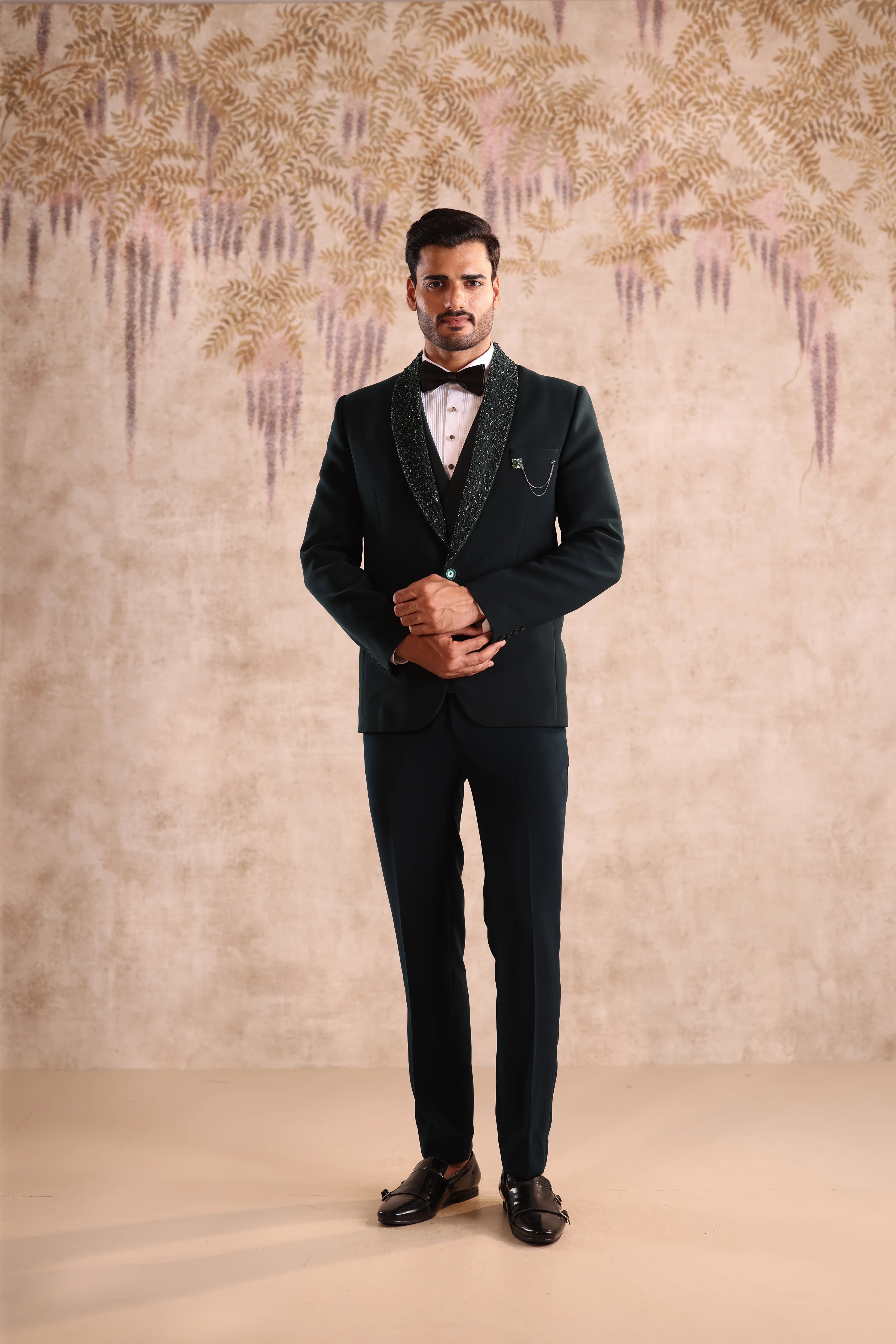 Man in a black suit standing against a decorative wall.