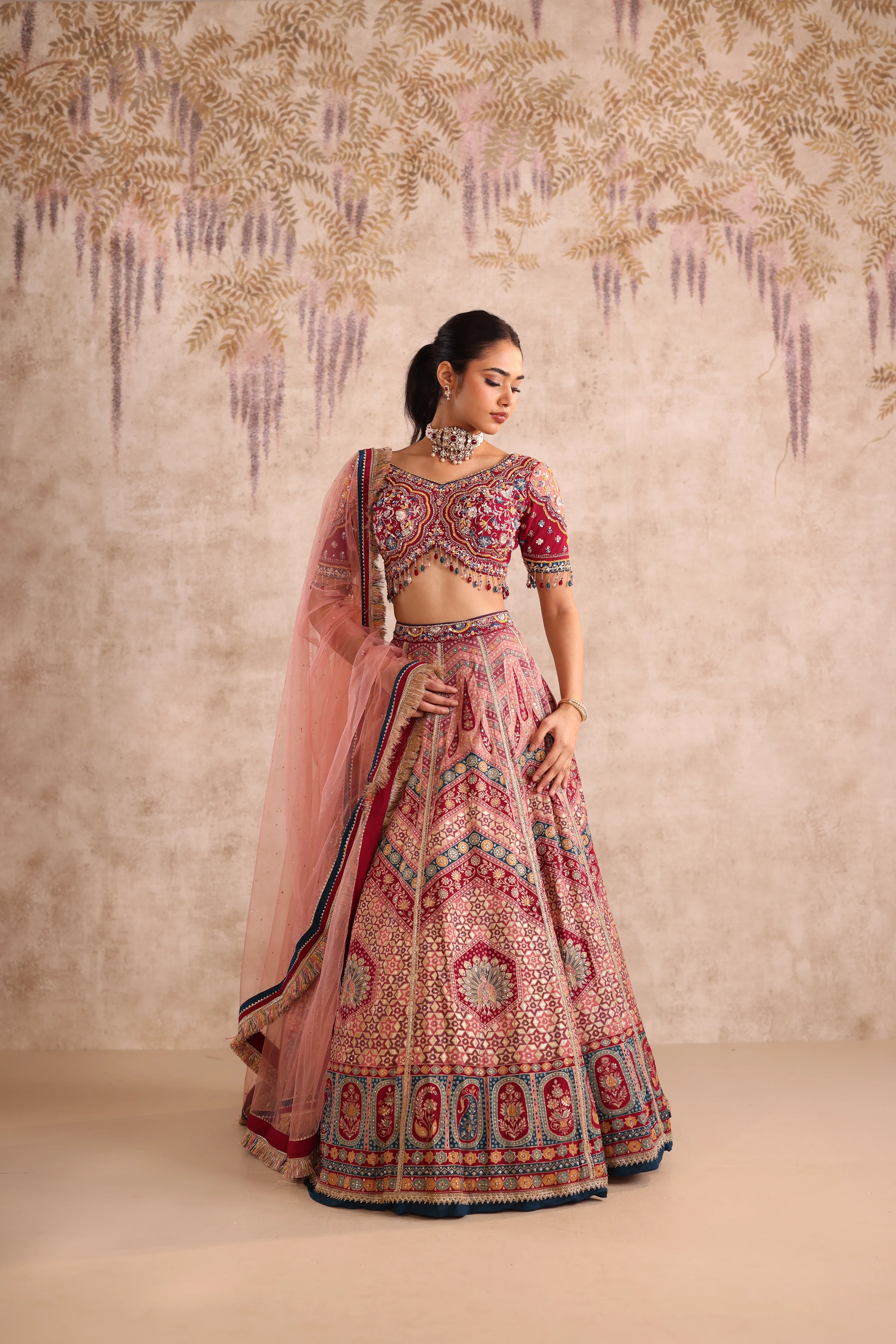 Woman in a traditional pink and red embroidered outfit against a decorative wall.