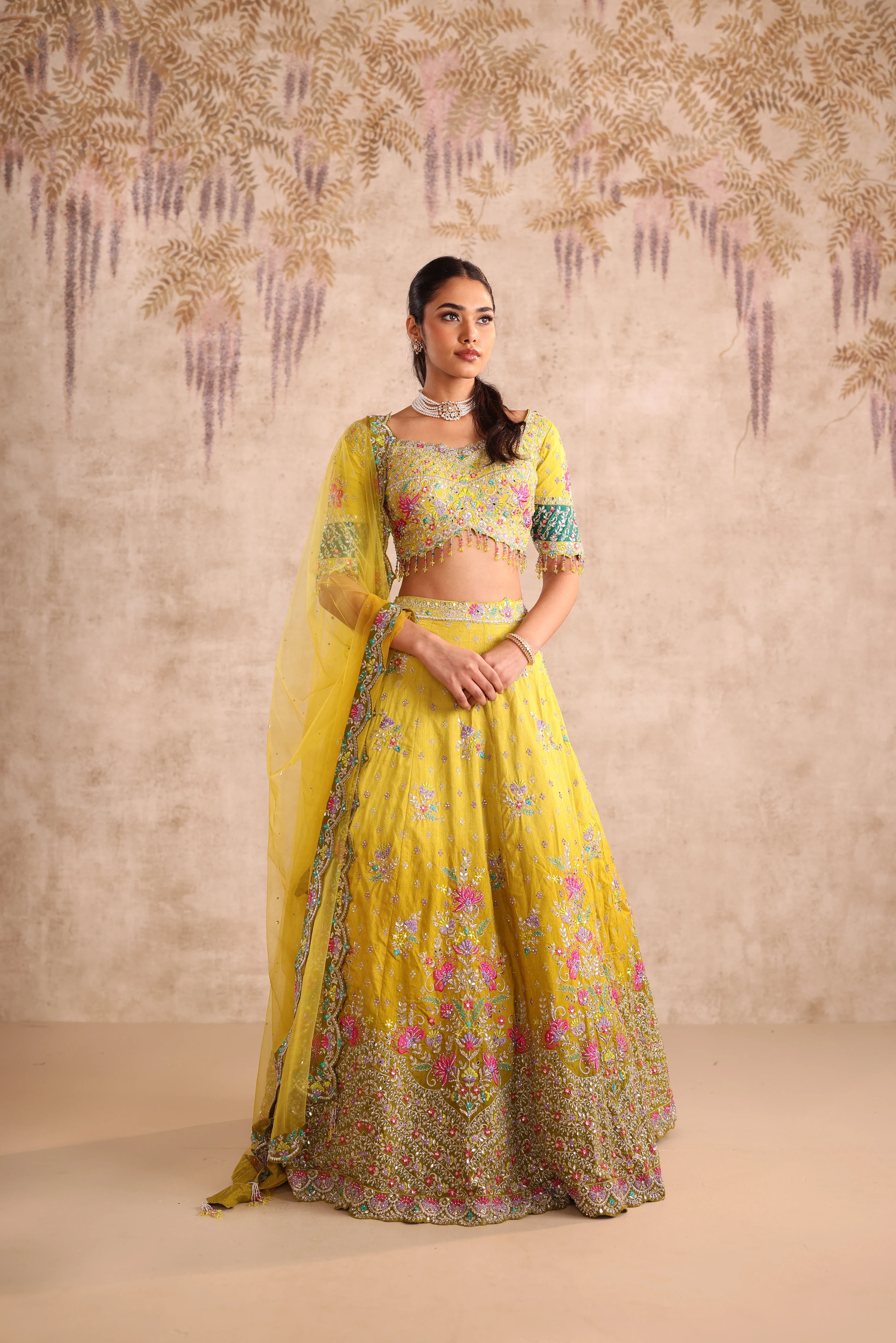 Woman in a yellow traditional outfit with floral patterns against a decorative wall.