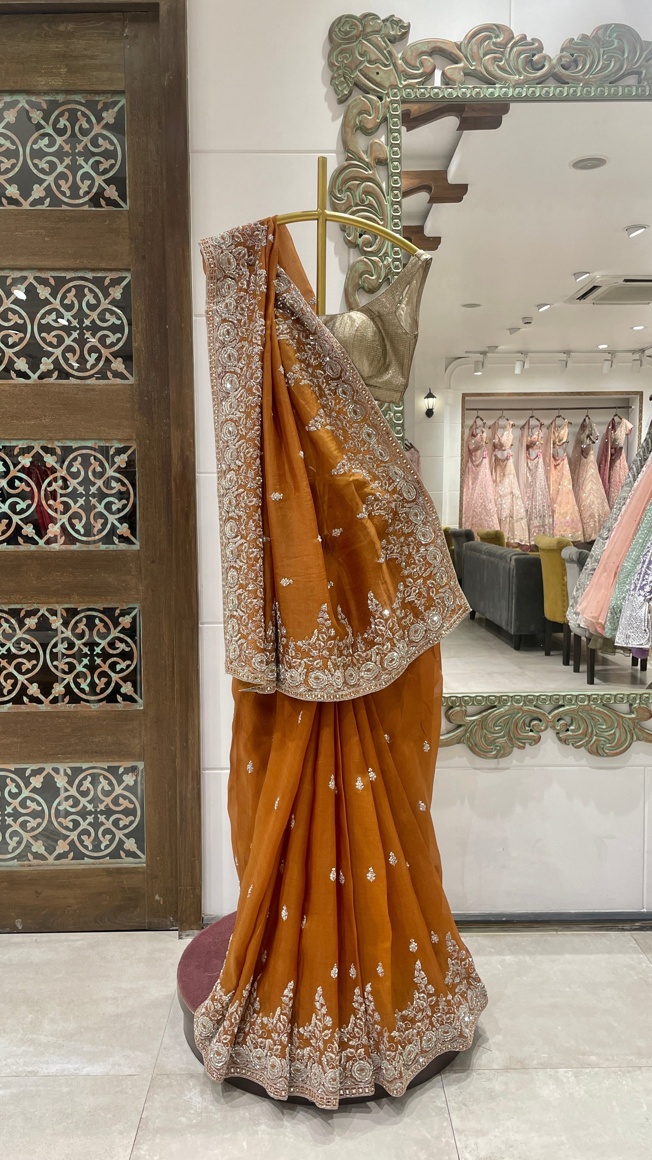 A rust-colored organza saree with zariwork displayed on a mannequin in a store with intricate zari work and a decorative border.