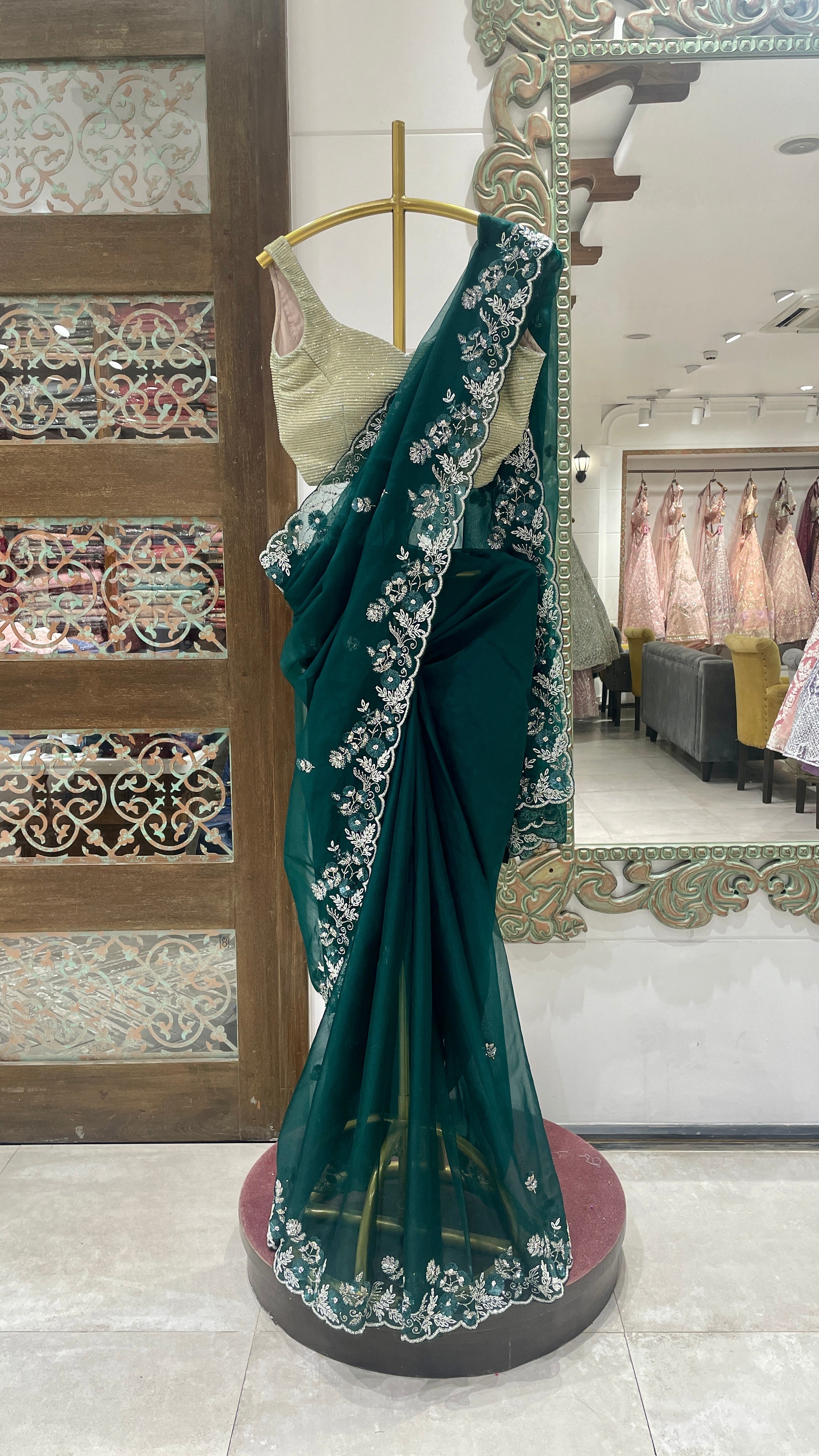 A green organza saree with sequins embellishments, draped over a mannequin, displayed in a retail setting with decorative patterns in the background.