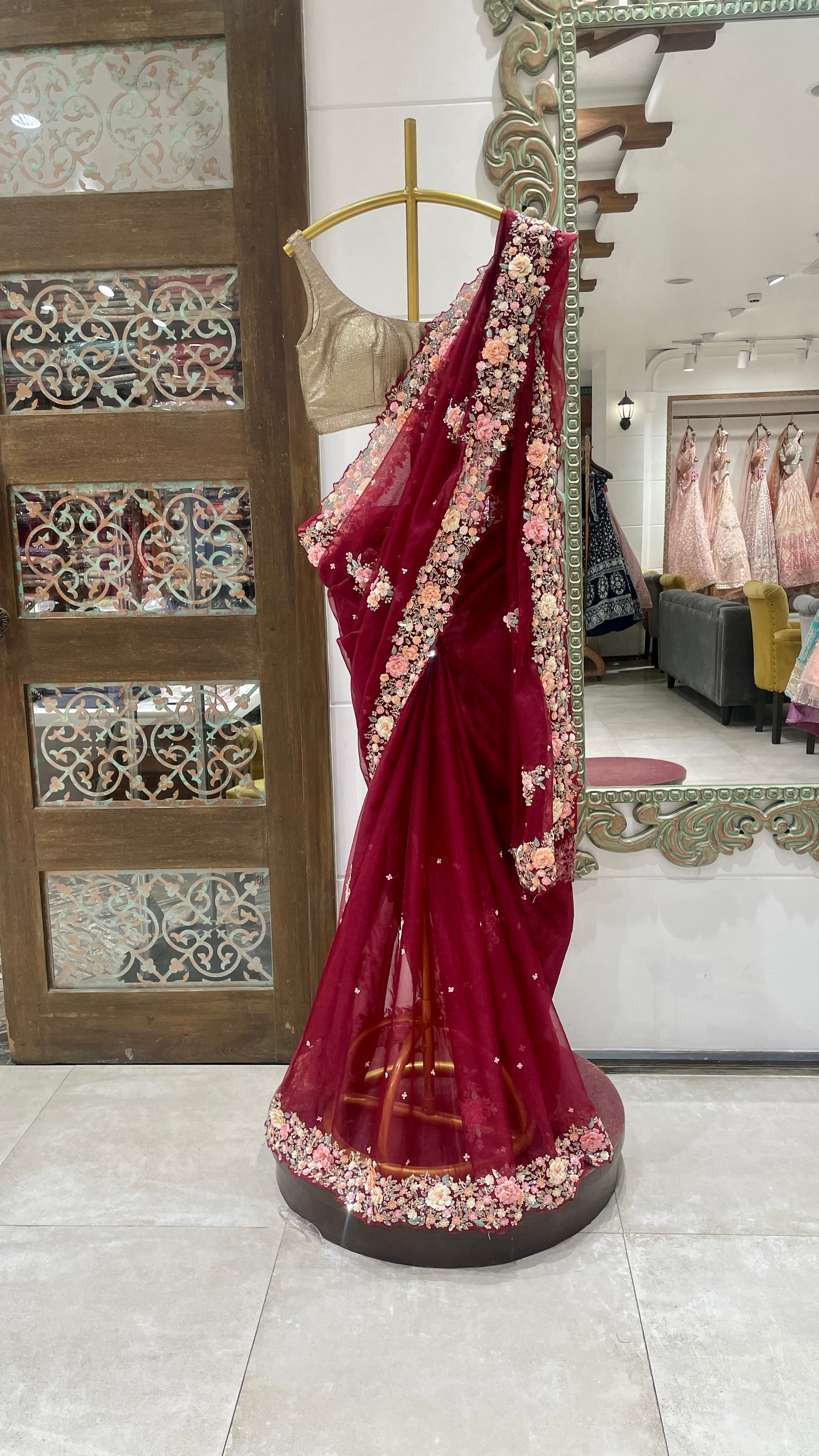 A red organza saree with embellishments displayed on a mannequin in a store with decorative patterns in the background.