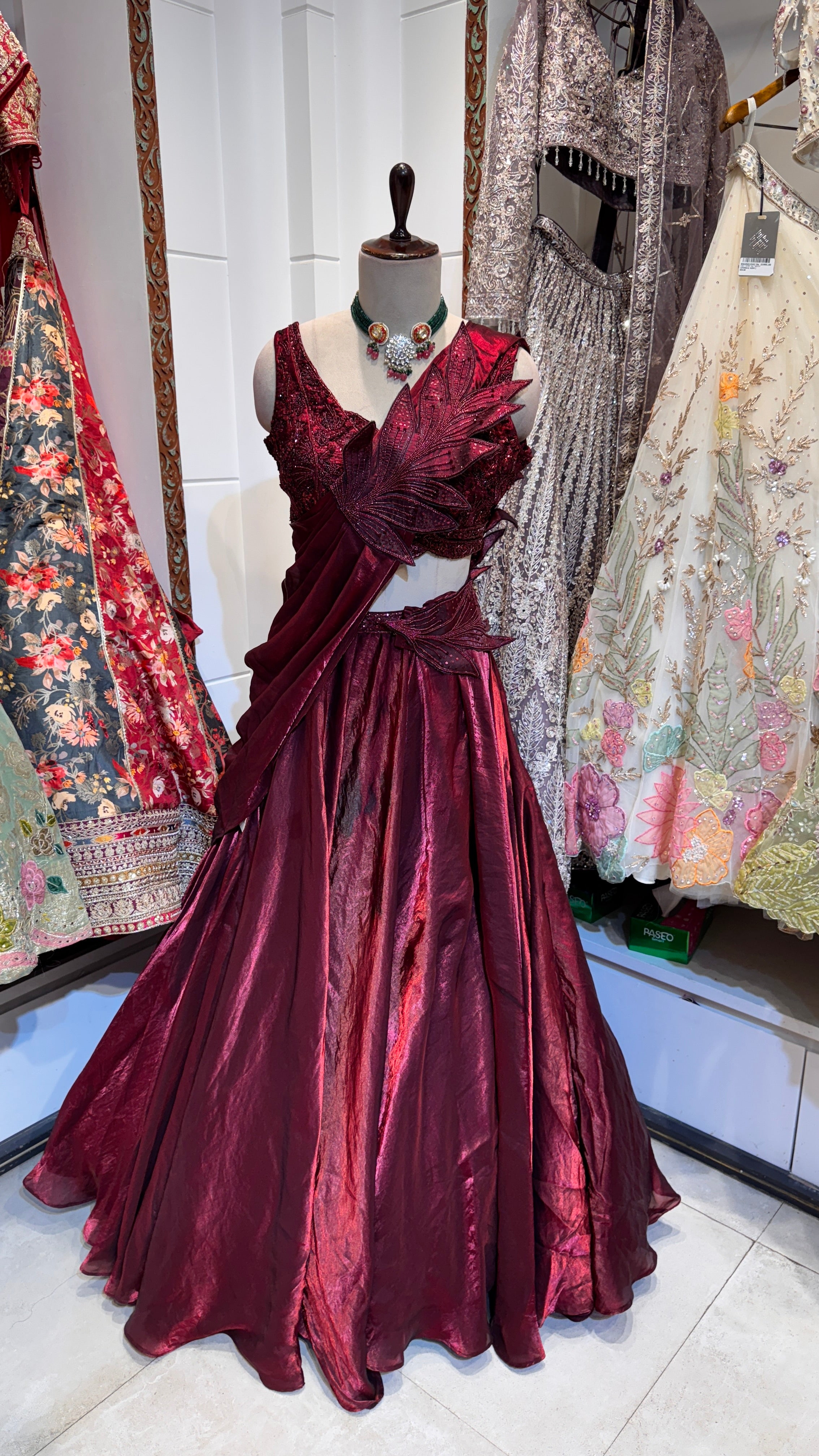 A red lehenga styled in a saree-draped silhouette with intricate embroidery and a matching embroidered blouse, displayed in a store setting.