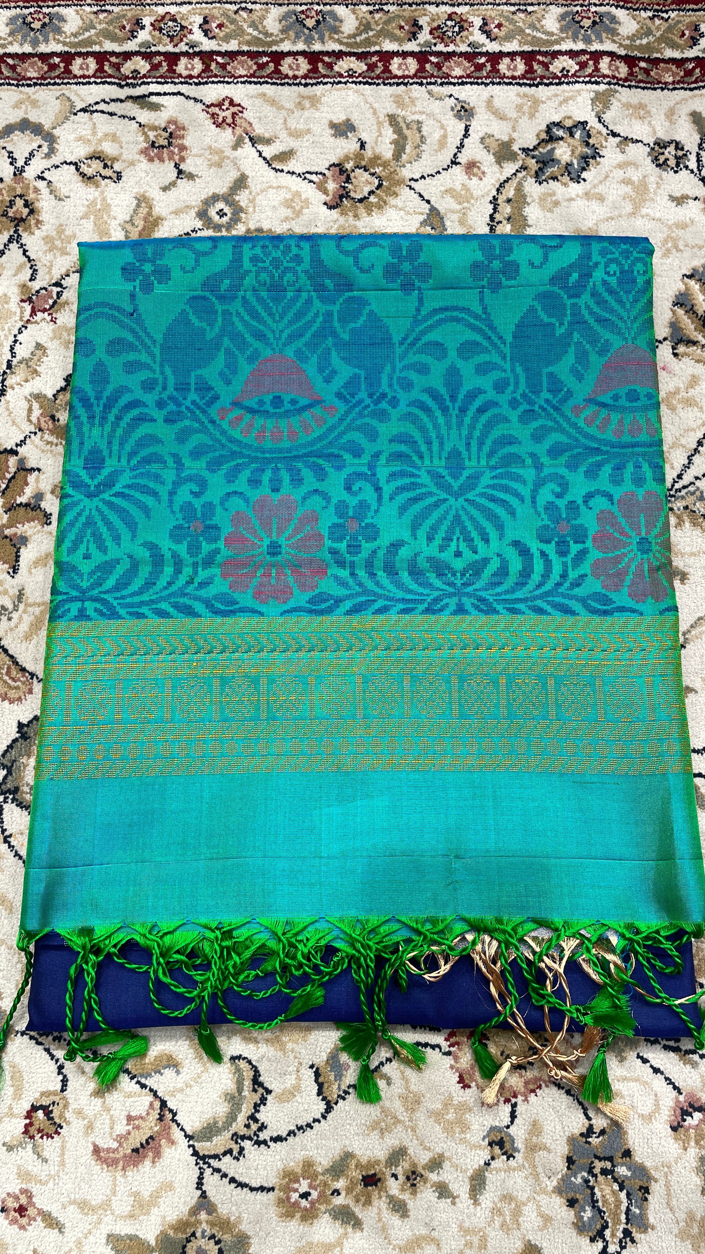 A folded turquoise blue handloom silk saree with intricate floral designs and a green border, displayed on a floral patterned fabric.