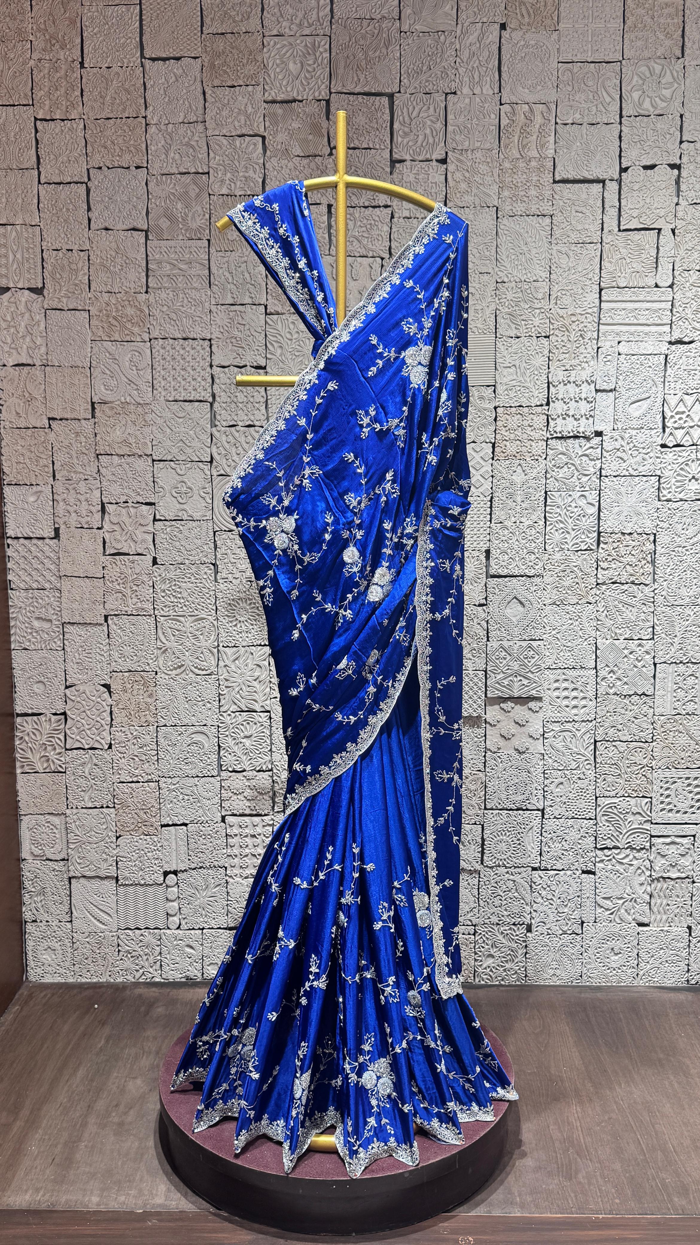 A blue saree with intricate silver embroidery displayed on a mannequin against a brick wall background.