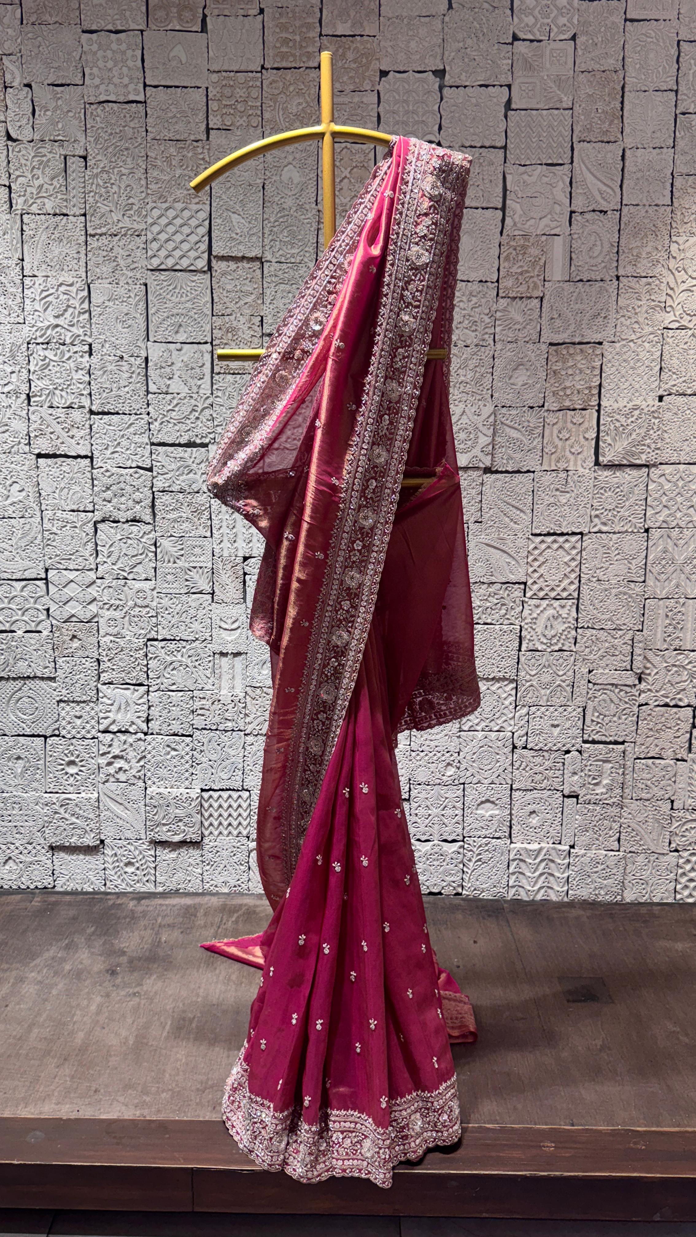 A tissue silk saree with heavy sequins border displayed on a hanger against a brick wall background, primarily pink in color with sequin embellishments.