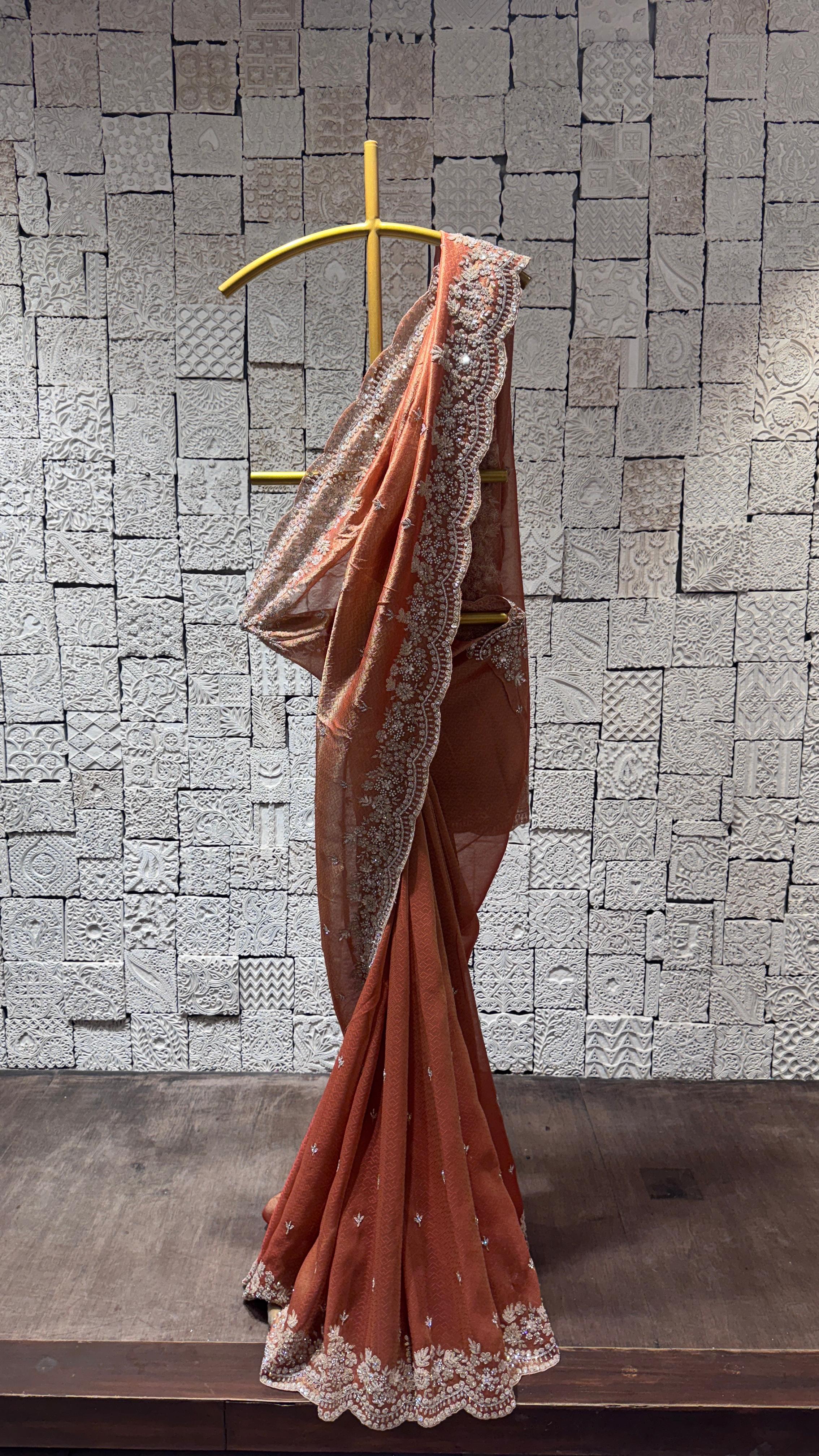 A peach-colored saree with pearl bead embroidery displayed on a hanger against a brick wall backdrop.