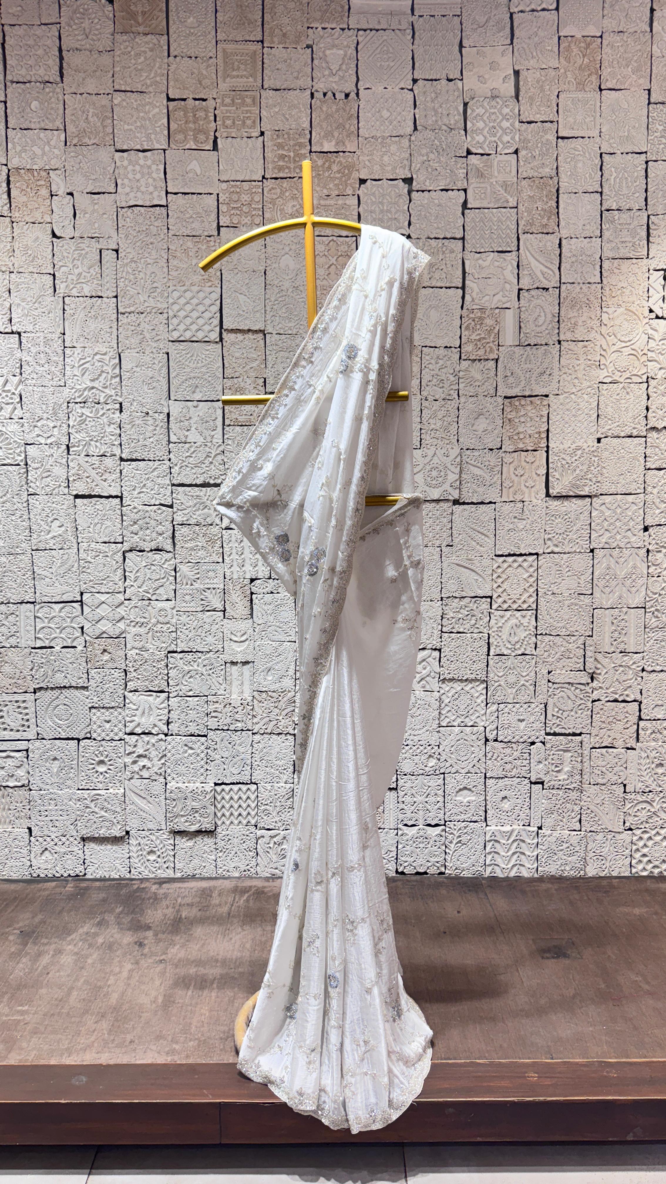 A white saree with intricate border and floral design displayed on a hanger against a brick wall backdrop.